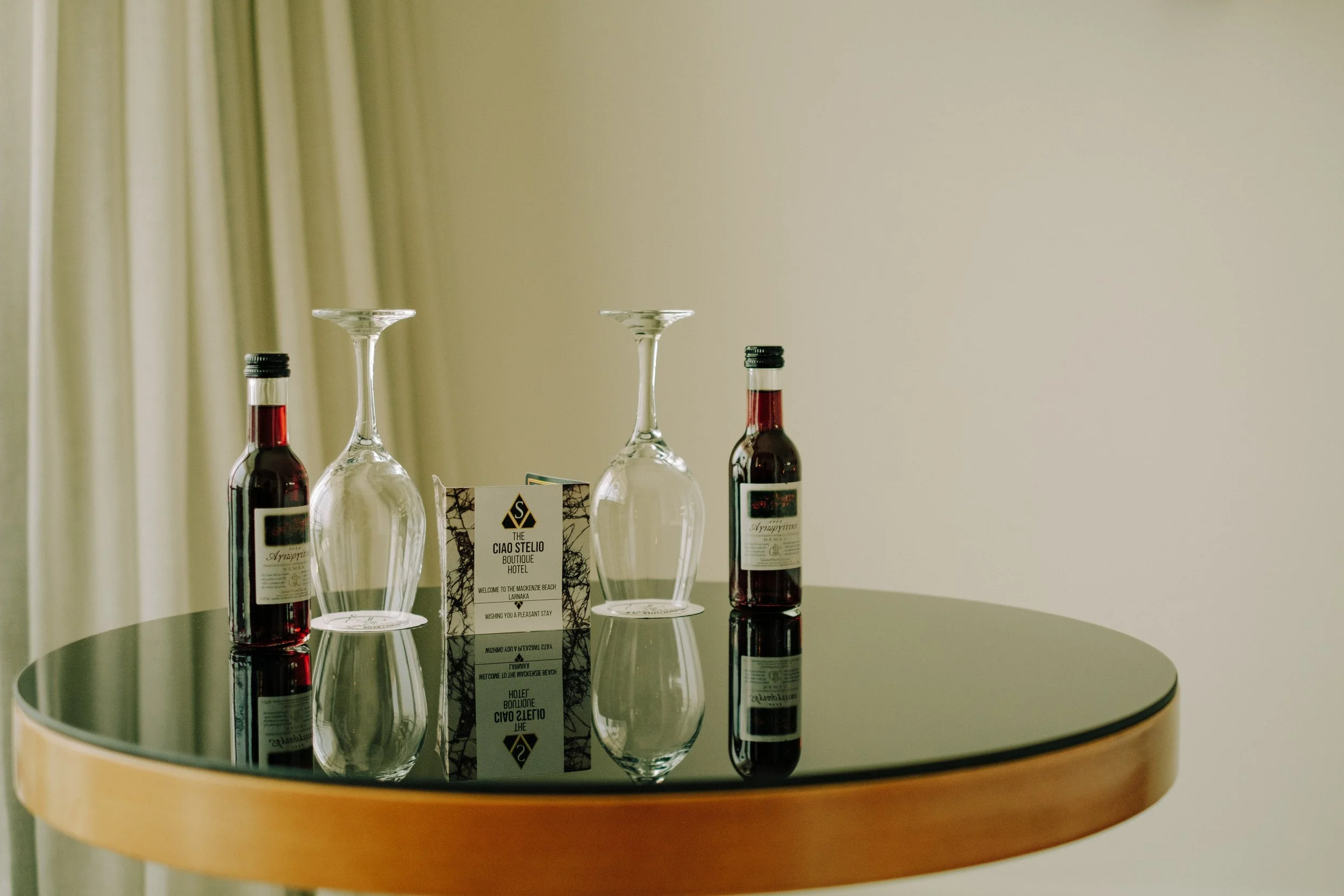 A round mirror table with two wine bottles, two upside-down glasses, and a hotel welcome card.