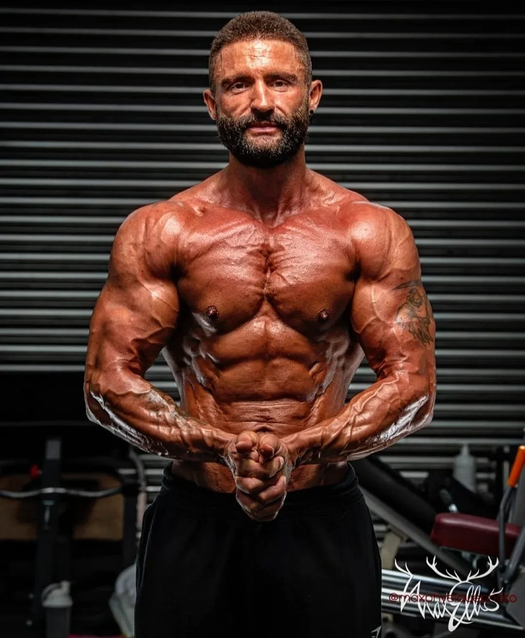 A muscular man with a beard and short hair, shirtless, flexing his arms and torso in a gym setting with black horizontal panels in the background.