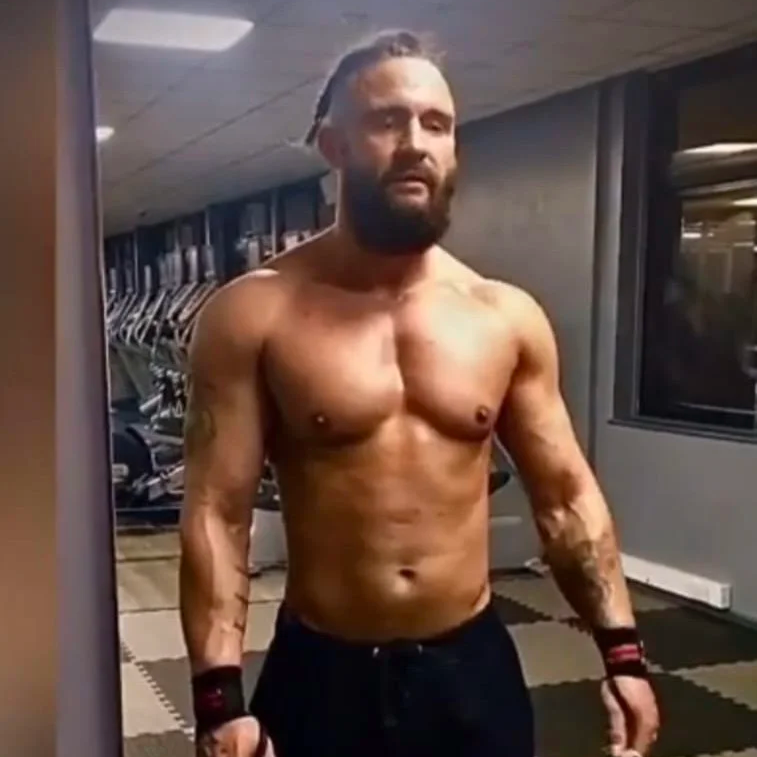 A shirtless man with tattoos and a beard taking a mirror selfie in a gym with exercise equipment in the background.