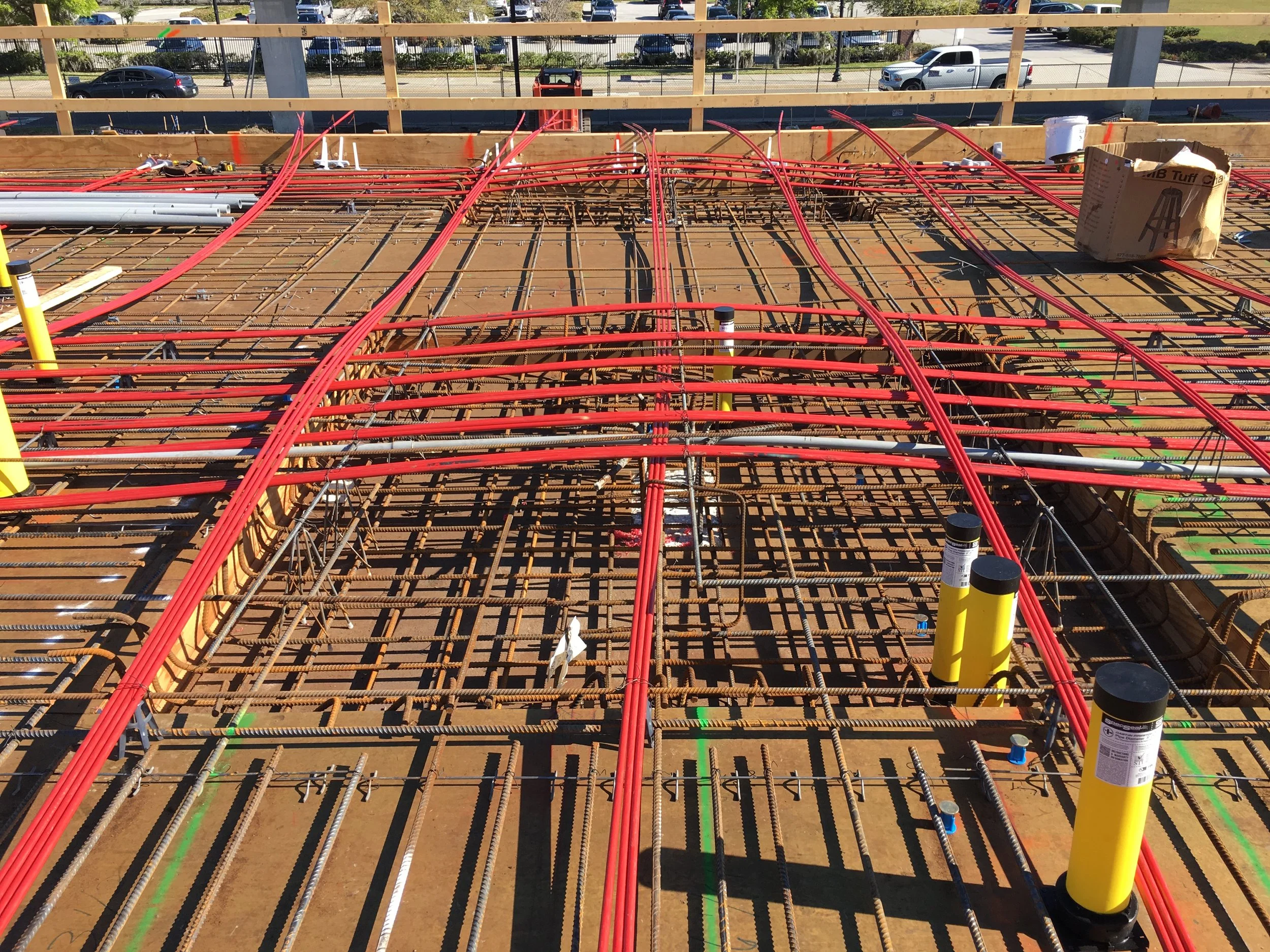 Construction site with exposed rebar and red plastic conduits for electrical wiring on a building's foundation. Yellow pipes are visible, and a cardboard box is in the background. Traffic and vehicles can be seen beyond the construction site.