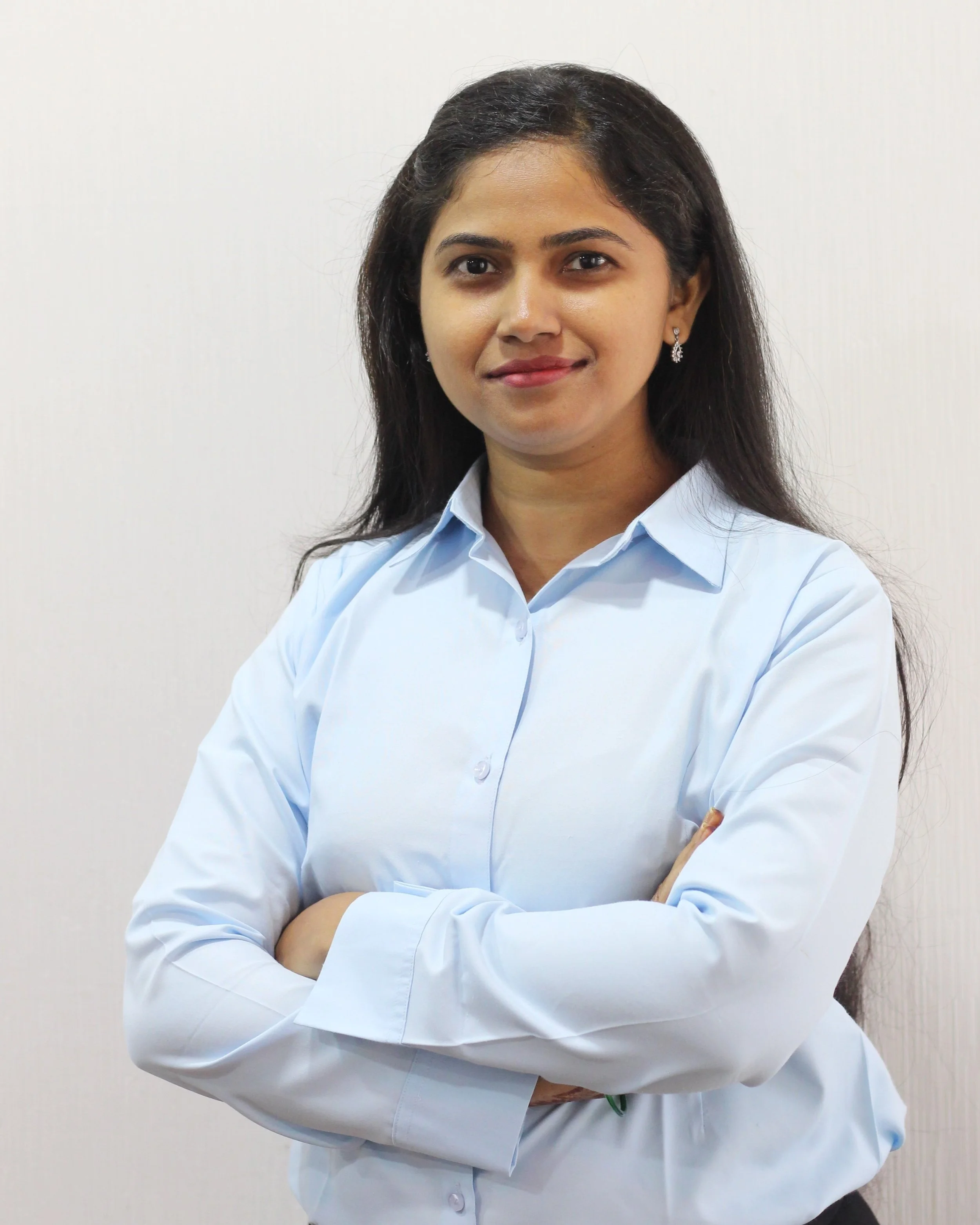 A woman with long black hair wearing a light blue button-up shirt, standing with arms crossed, smiling slightly, against a plain white background.