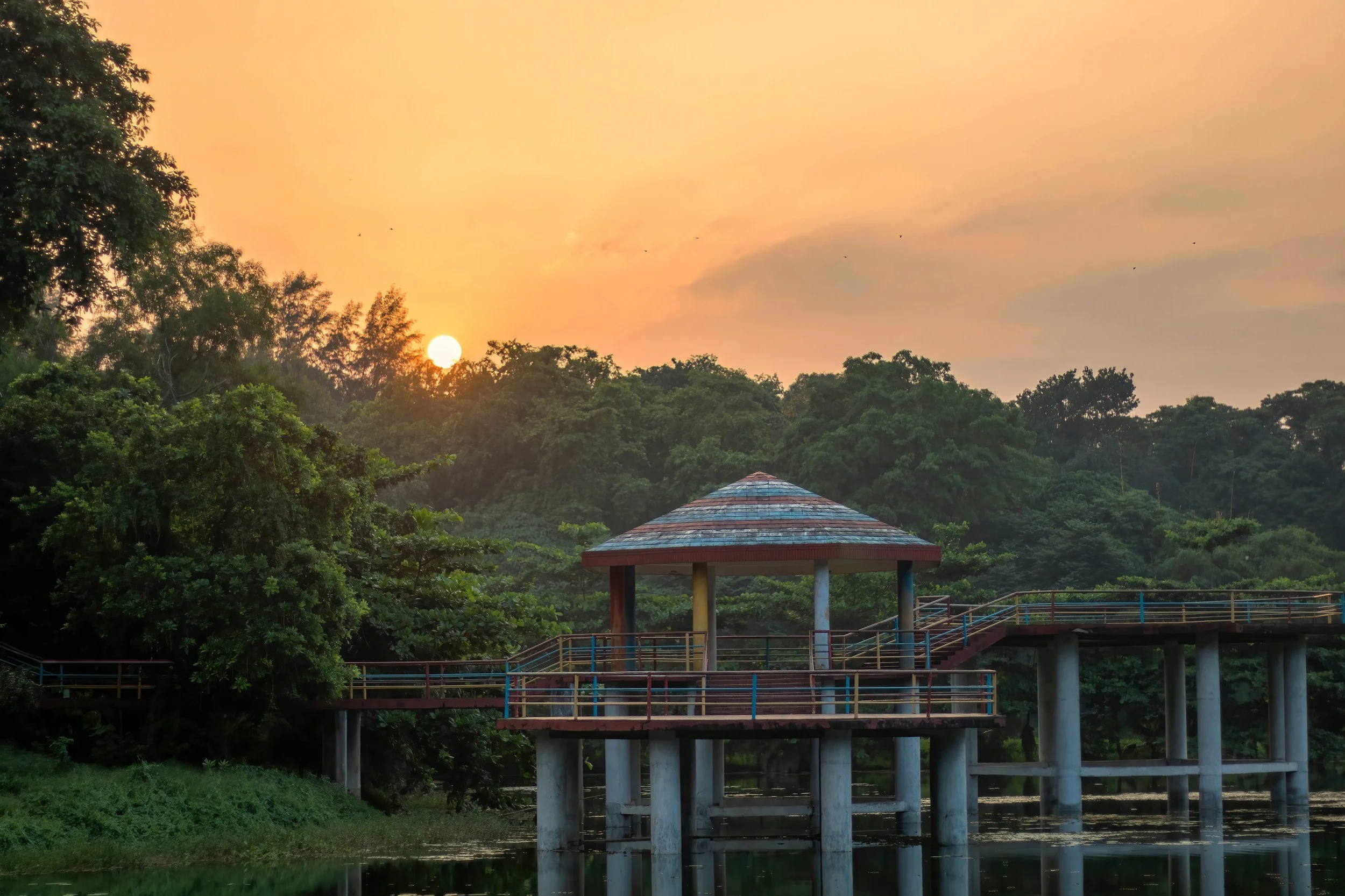 Sunset over a lush green park with a small pavilion on stilts over a lake, surrounded by trees.