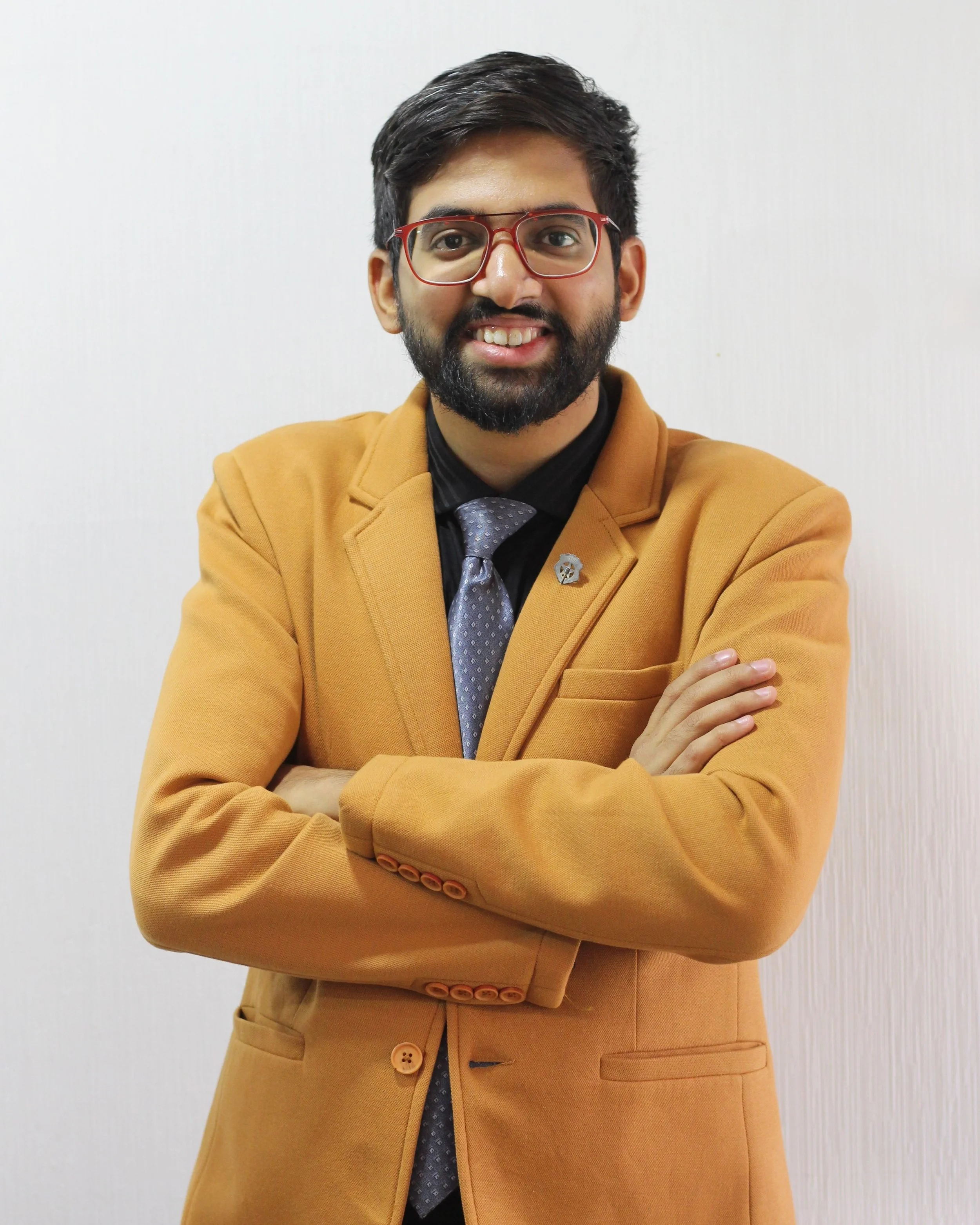 A man with glasses, beard, and dark hair standing against a plain light background, wearing a mustard-colored blazer, a black shirt, a patterned gray tie, and a lapel pin.