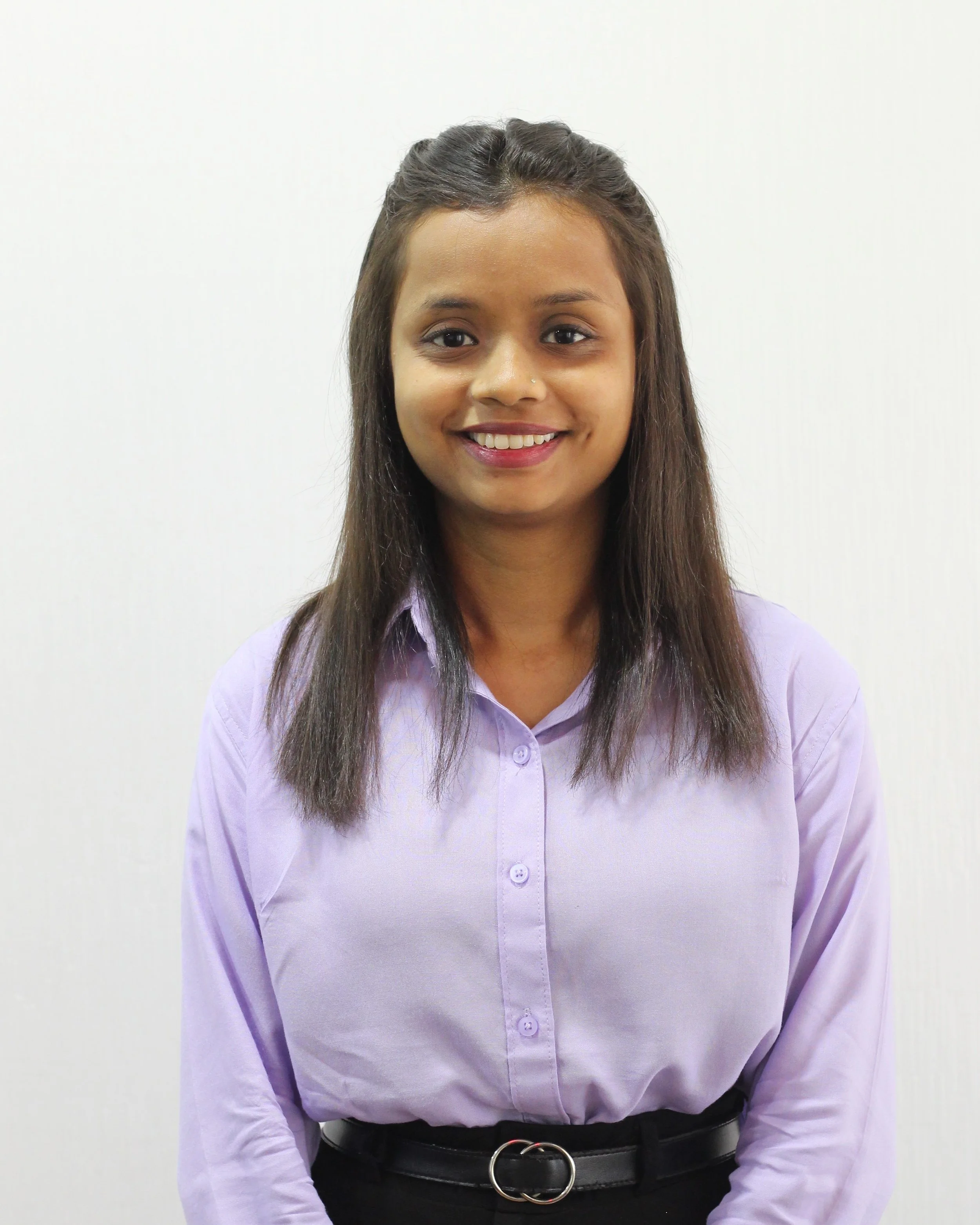 A young woman with long dark hair, smiling, wearing a light purple button-up shirt and a black belt with a circular buckle, standing against a plain white background.