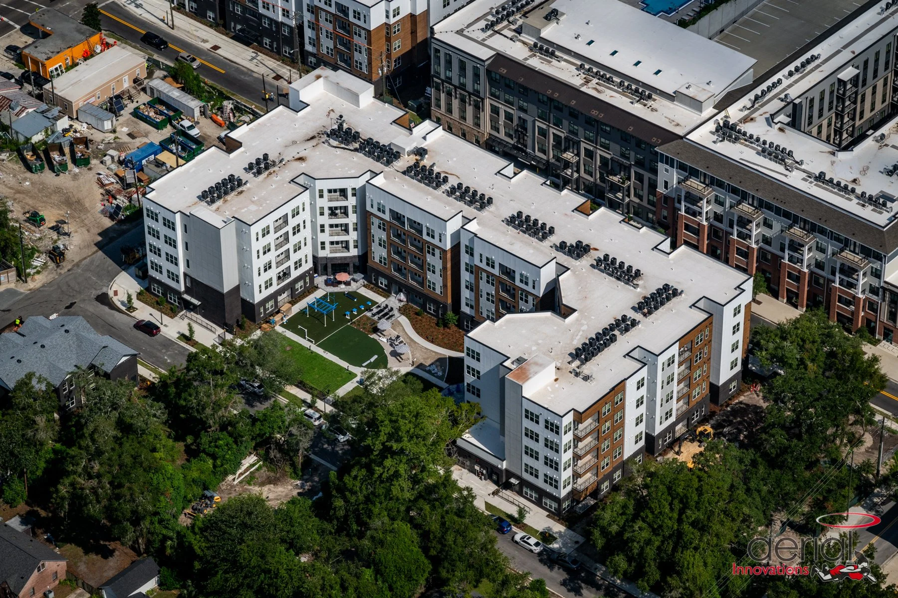 Aerial view of modern apartment complex with white, brown, and black exterior, rooftop HVAC units, green courtyard with playground, and surrounding trees and streets.