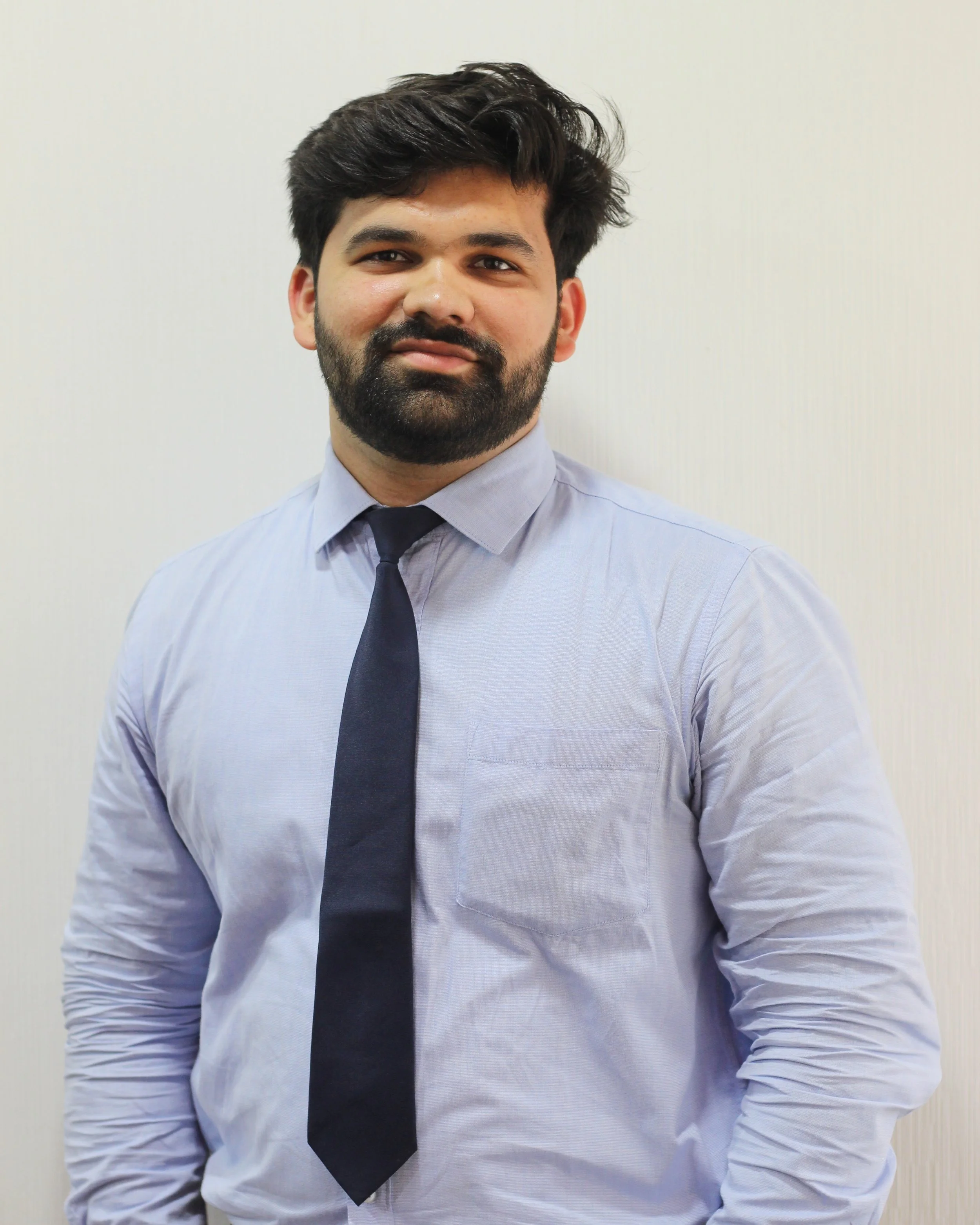 A man with dark hair and a beard wearing a light blue dress shirt and a dark tie, standing against a plain white wall.
