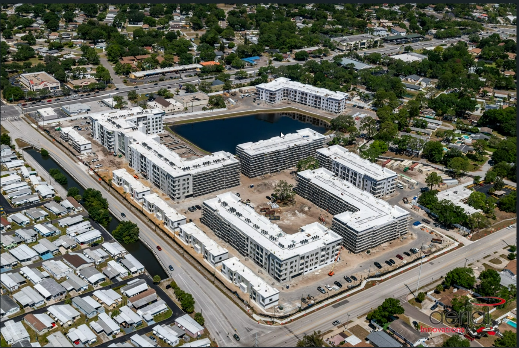 Aerial view of a residential construction site with multiple modern apartment buildings and a pond in the center, surrounded by a mix of commercial and residential neighborhoods.
