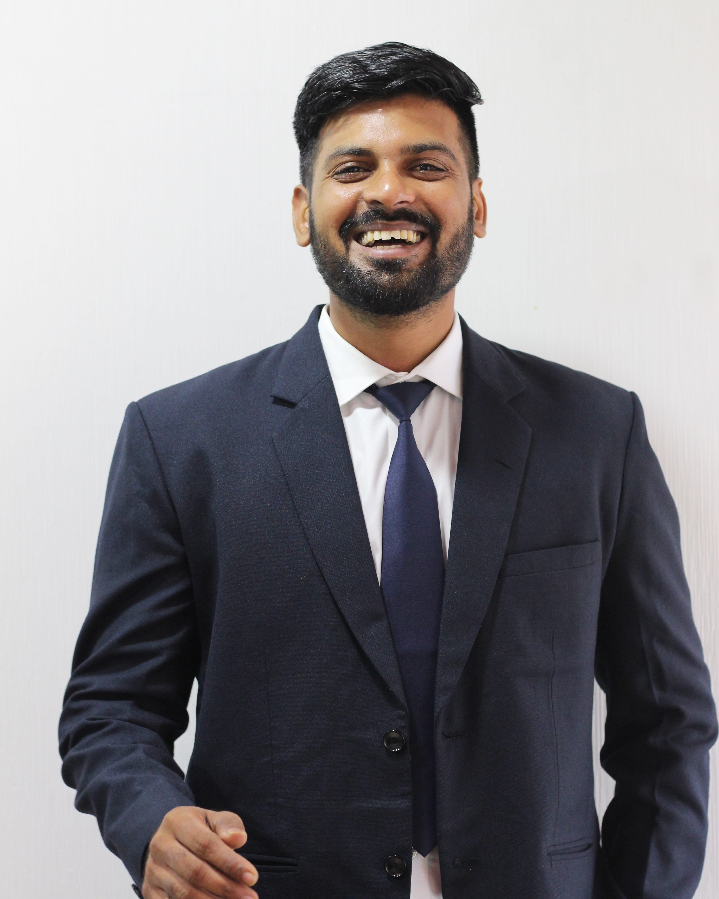 A man in a black suit, white shirt, and navy tie smiling against a plain white background.