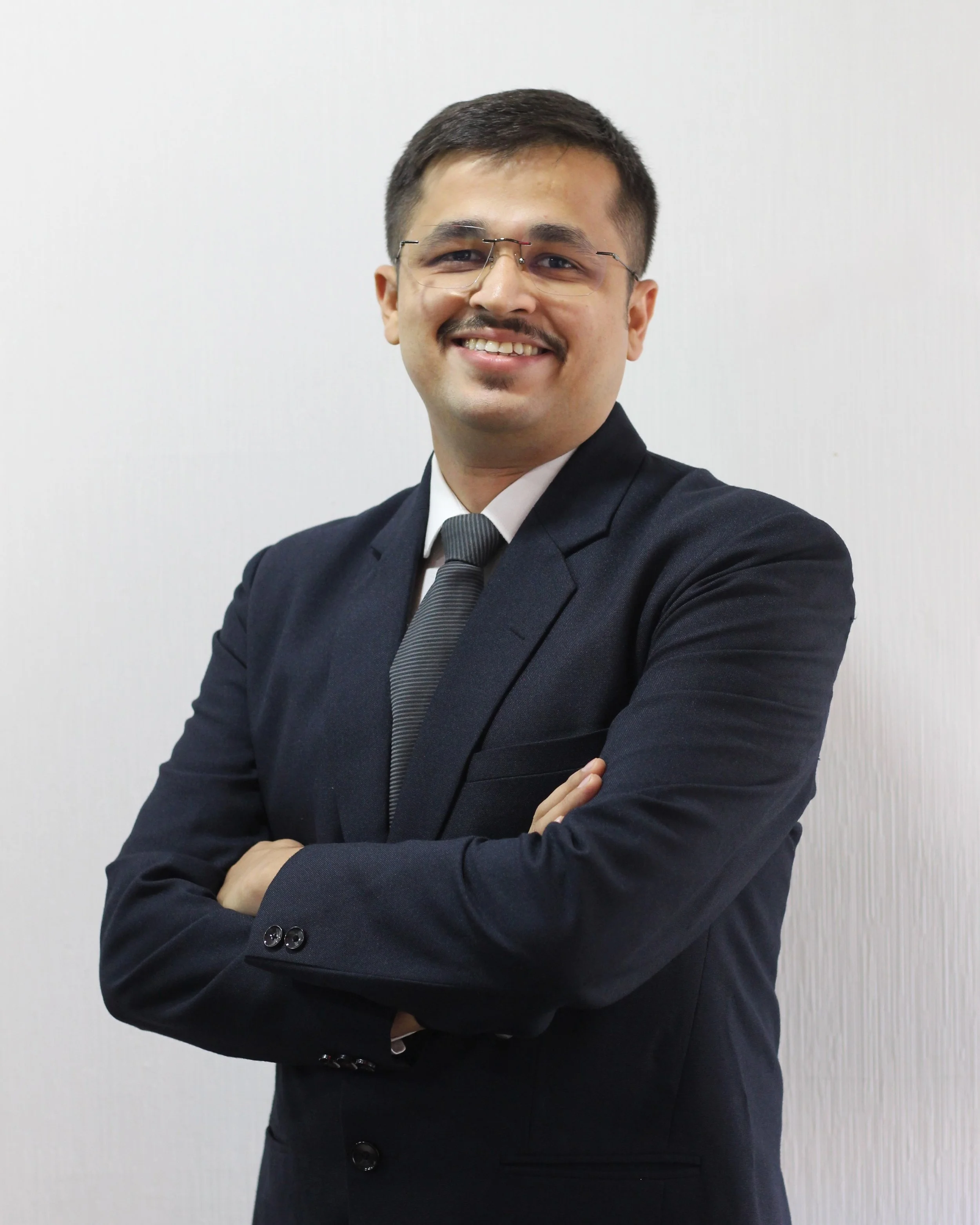 A man in a dark suit, white shirt, and glasses with arms crossed, smiling, standing against a plain, light-colored wall.