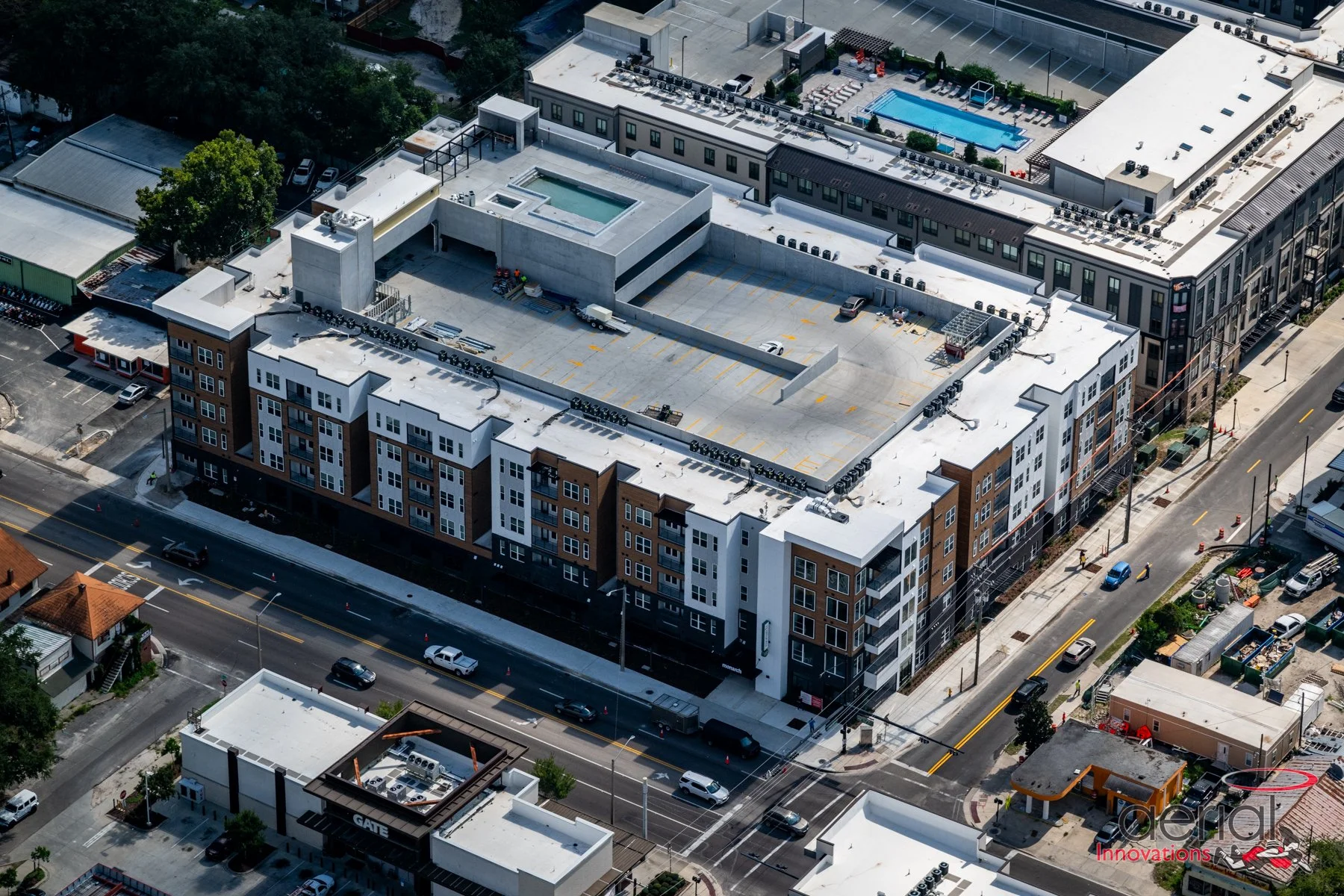Aerial view of a modern multi-story apartment complex with a rooftop pool and parking lot, surrounded by streets and other buildings.