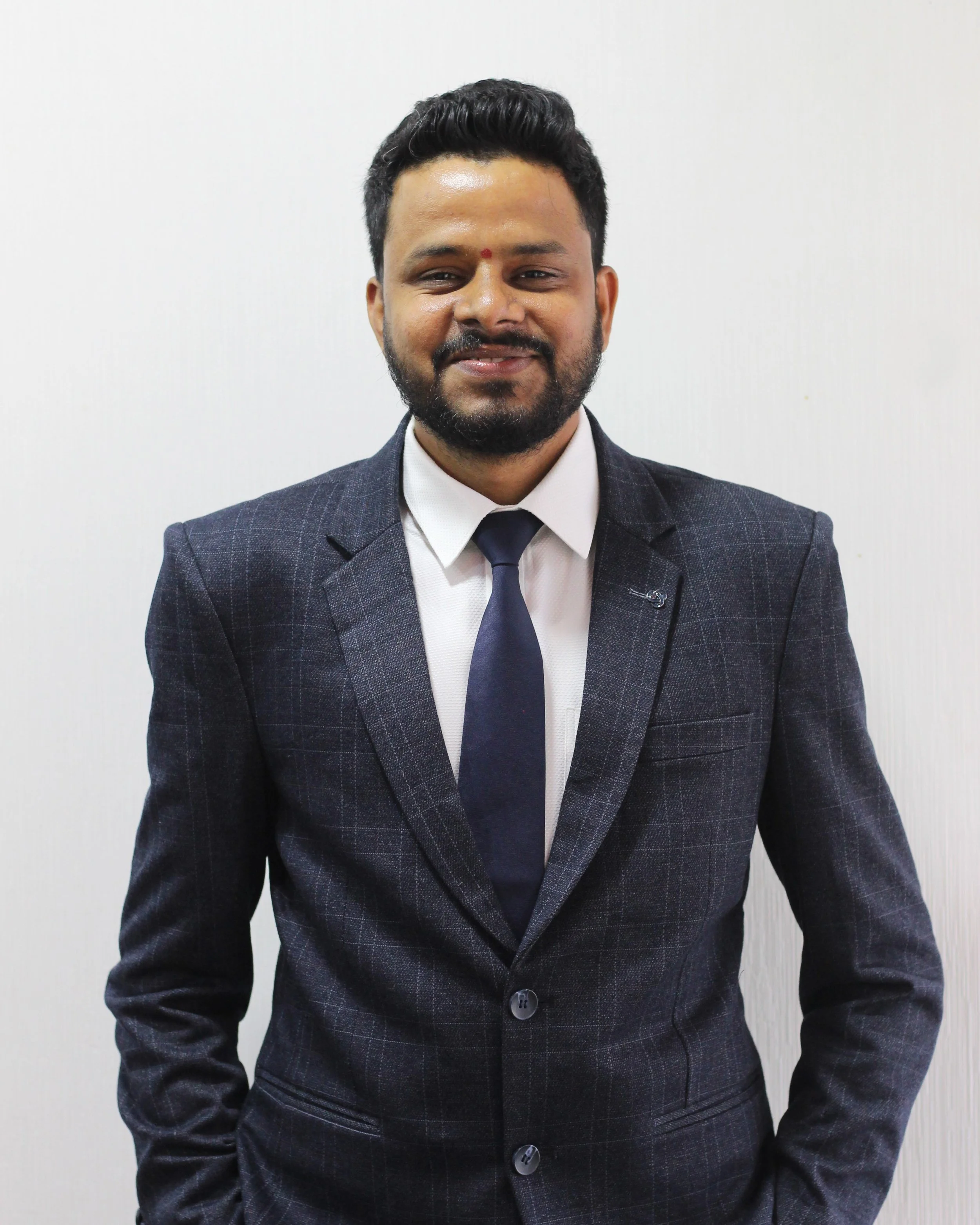 Man in a dark gray suit, white shirt, and navy tie posing for a photo against a plain white wall.