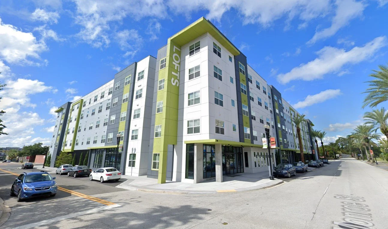 Modern multi-story apartment building with green accents and a sign that reads 'LOFTS' on the corner, with cars parked along the street and palm trees lining the sidewalk.