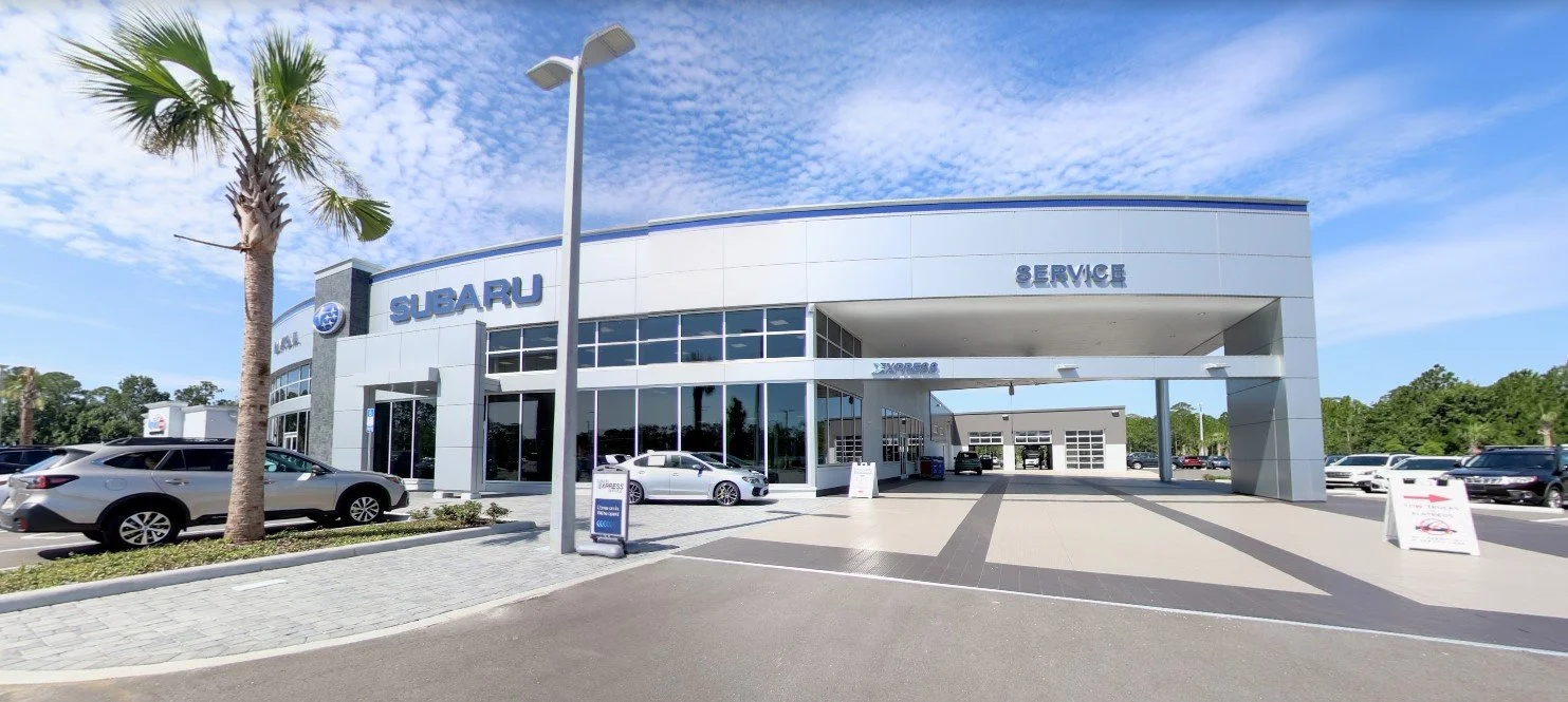 Exterior of a Subaru car dealership with cars parked outside, a blue sky with clouds, and palm trees.