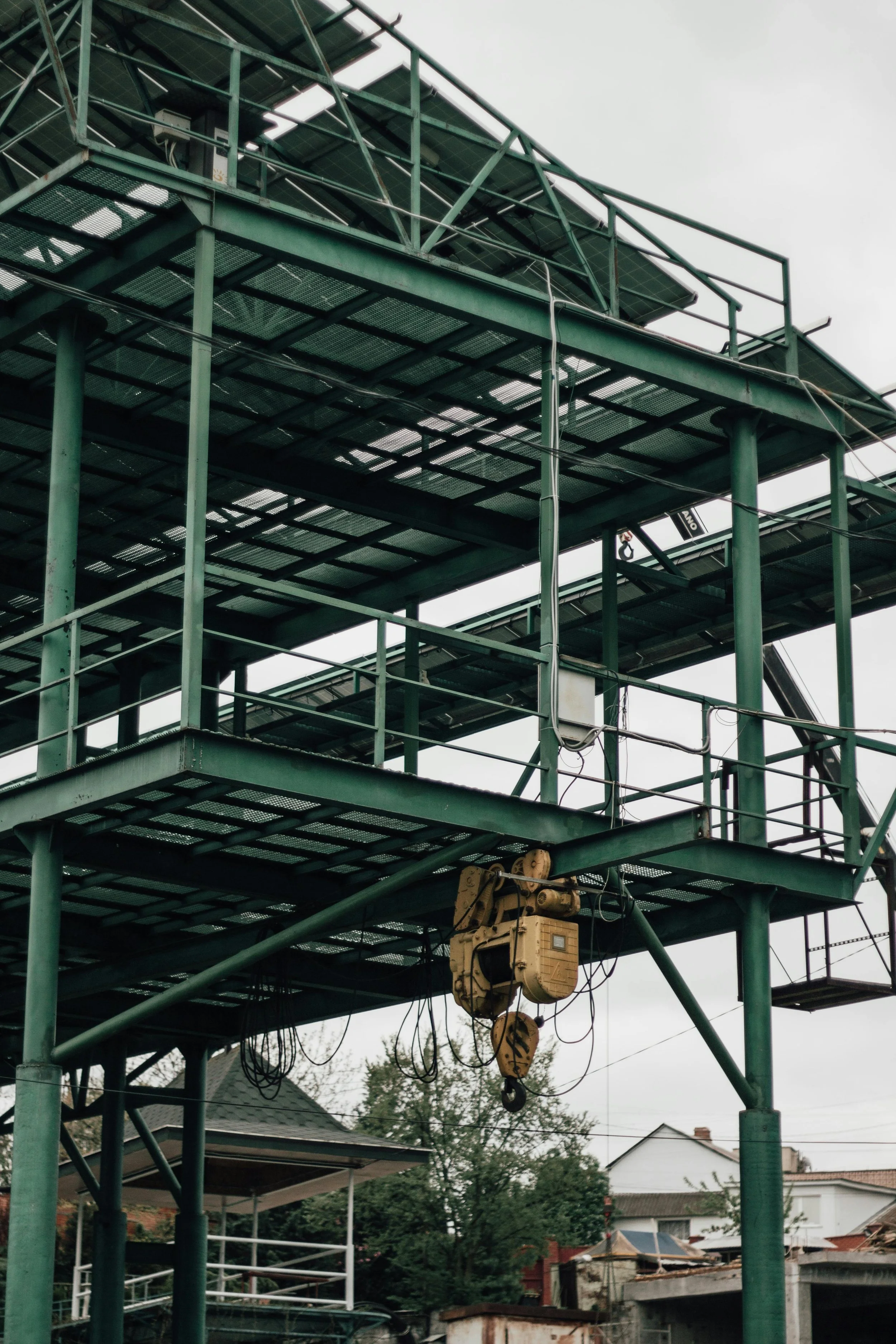 Green metal scaffolding structure with stairs and railings, surrounding a construction or maintenance area, with residential houses and trees in the background under an overcast sky.