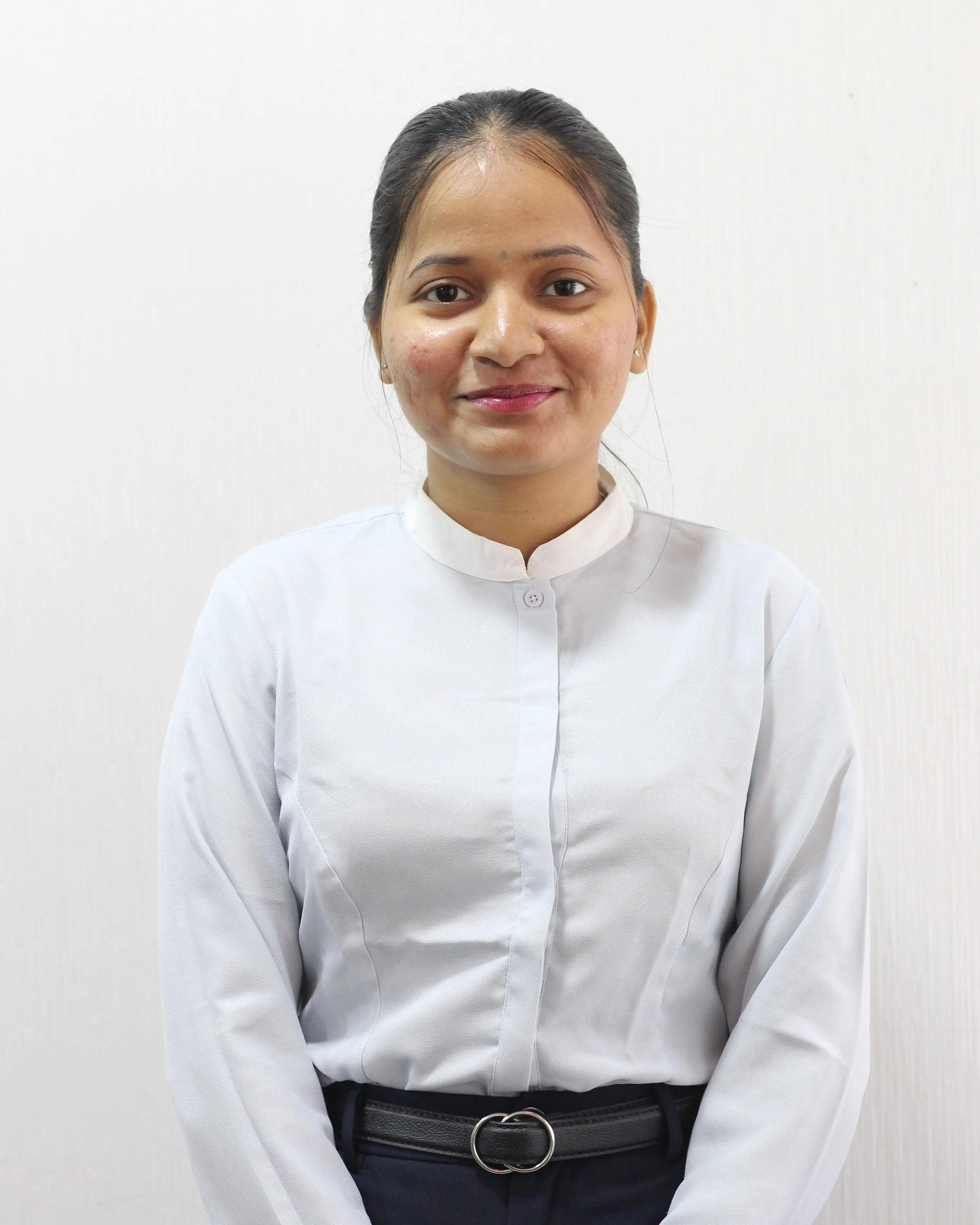 A woman with dark hair tied back, wearing a white collared shirt, standing against a plain white wall.