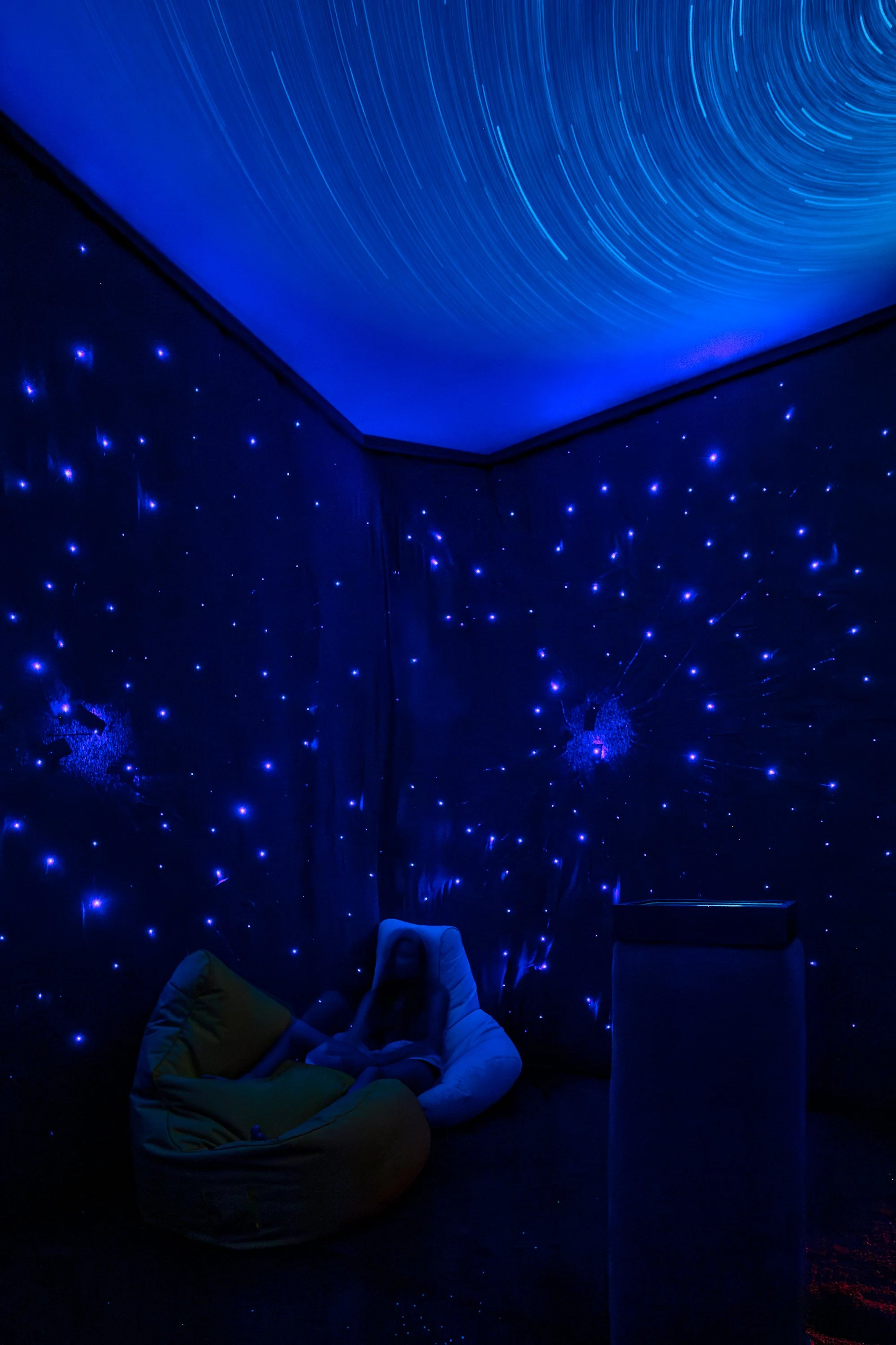 A person relaxing on a bean bag chair in a dark room with star constellation projections on the walls and ceiling, illuminated by blue lights simulating stars.