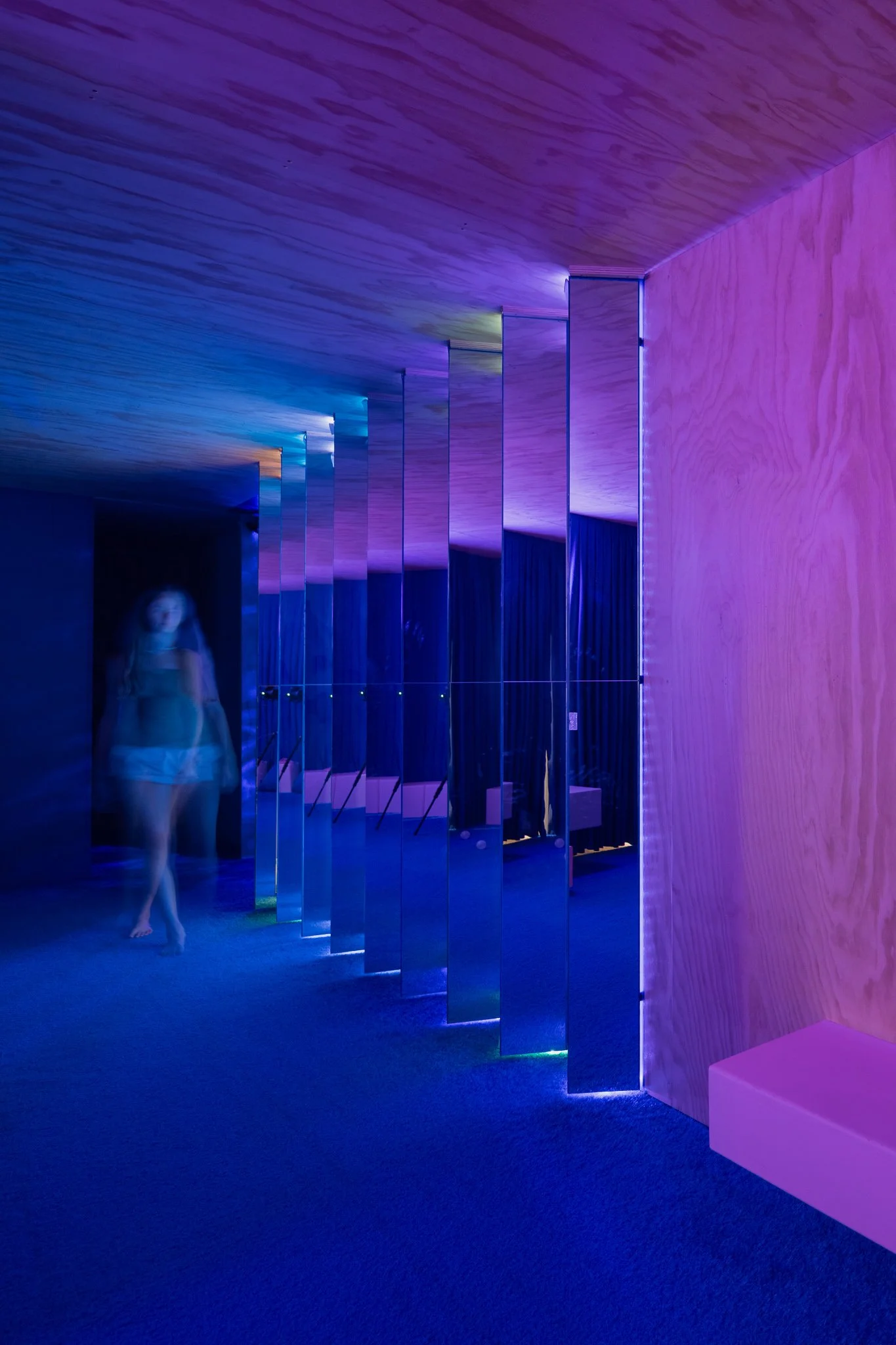 A hallway with mirrors on one side, illuminated by colored LED lights creating a rainbow effect on the ceiling. A blurred figure of a woman stands near the mirrors.