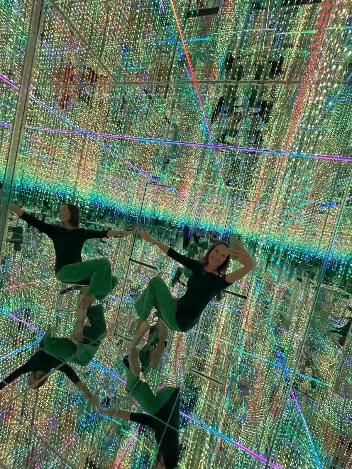 A woman in black top and green pants taking a selfie inside a mirrored room filled with colorful, shimmering LED lights. The room creates a kaleidoscopic effect with reflections.
