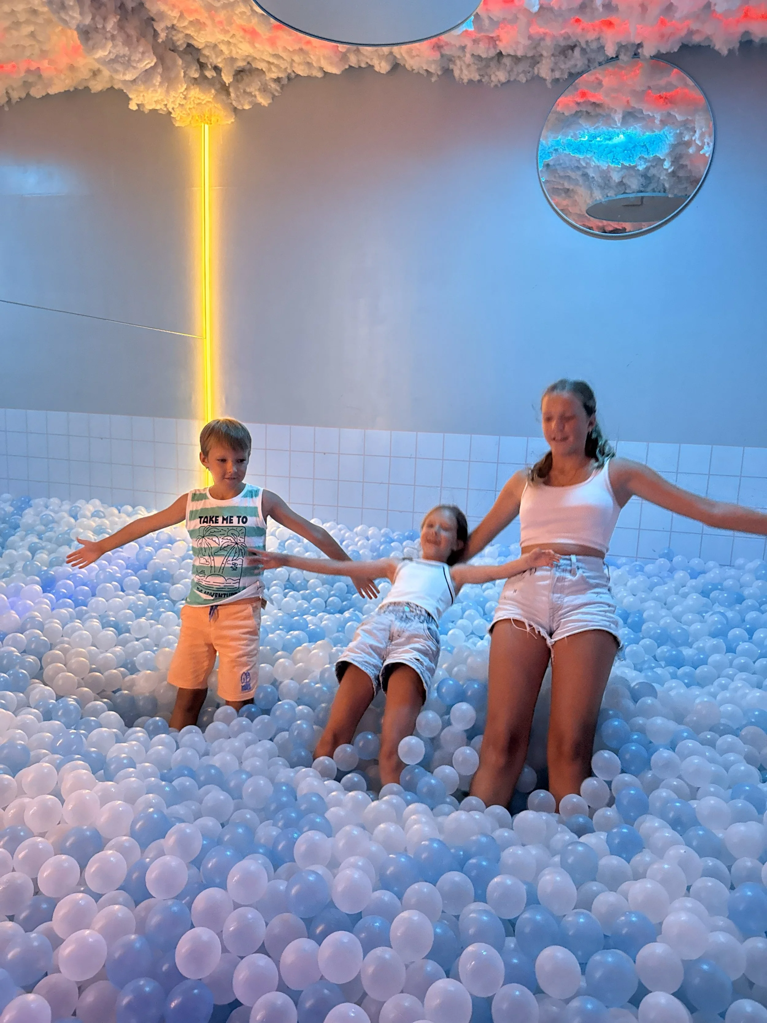 Three children are playing in a ball pit filled with white plastic balls in an indoor play area. The background features colorful lighting and ceiling decorations resembling clouds.
