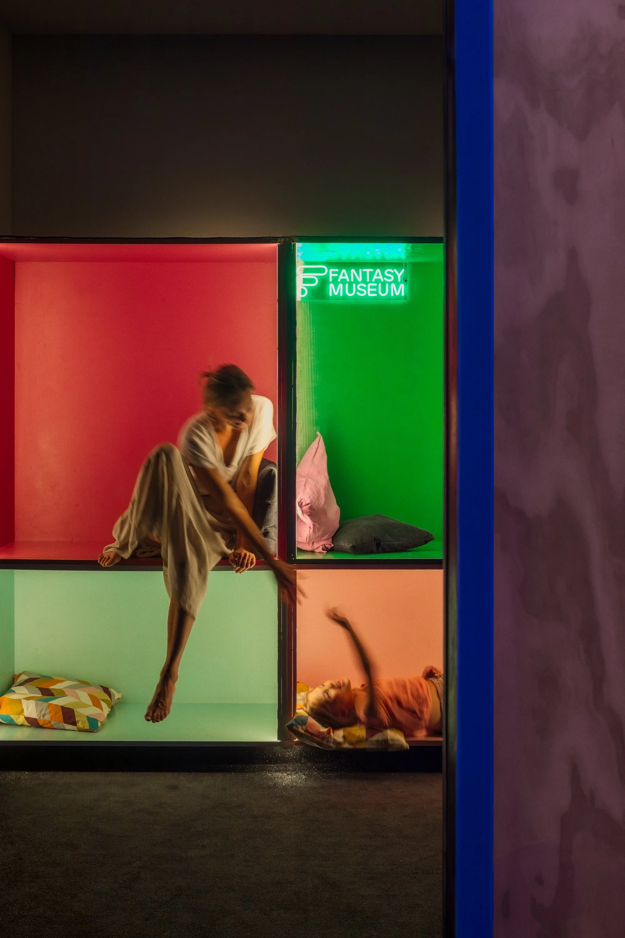 A person in beige pajamas sitting on the edge of a colorful cubby wall, with one leg hanging down and the other bent, inside a neon-lit room labeled 'FANTASY MUSEUM'. Two children are lying inside the cubby with colorful pillows, one waving their arm, and the other resting.