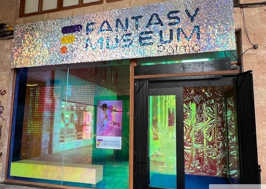 The exterior of the Fantasy Museum has a holographic, reflective façade with a large glass window. The sign above the entrance reads 'Fantasy Museum' in colorful, sparkly lettering. Inside, there are vibrant, colorful displays and a poster showing a person in a fantasy costume.