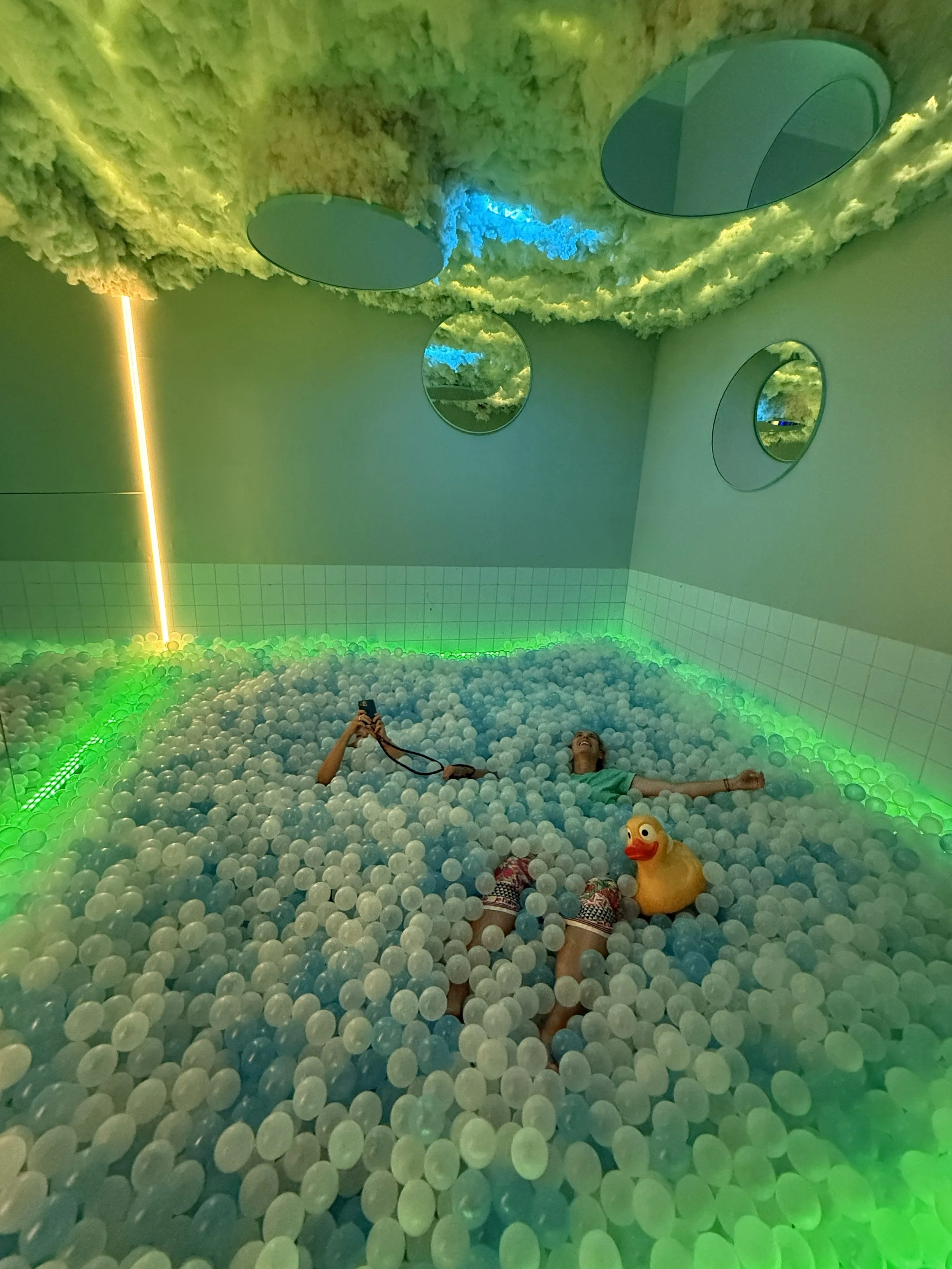 Person lying in a ball pit filled with white balls, with a rubber duck float, in a room with green and blue lighting, circular mirrors on the walls, and a cloud-like textured ceiling.