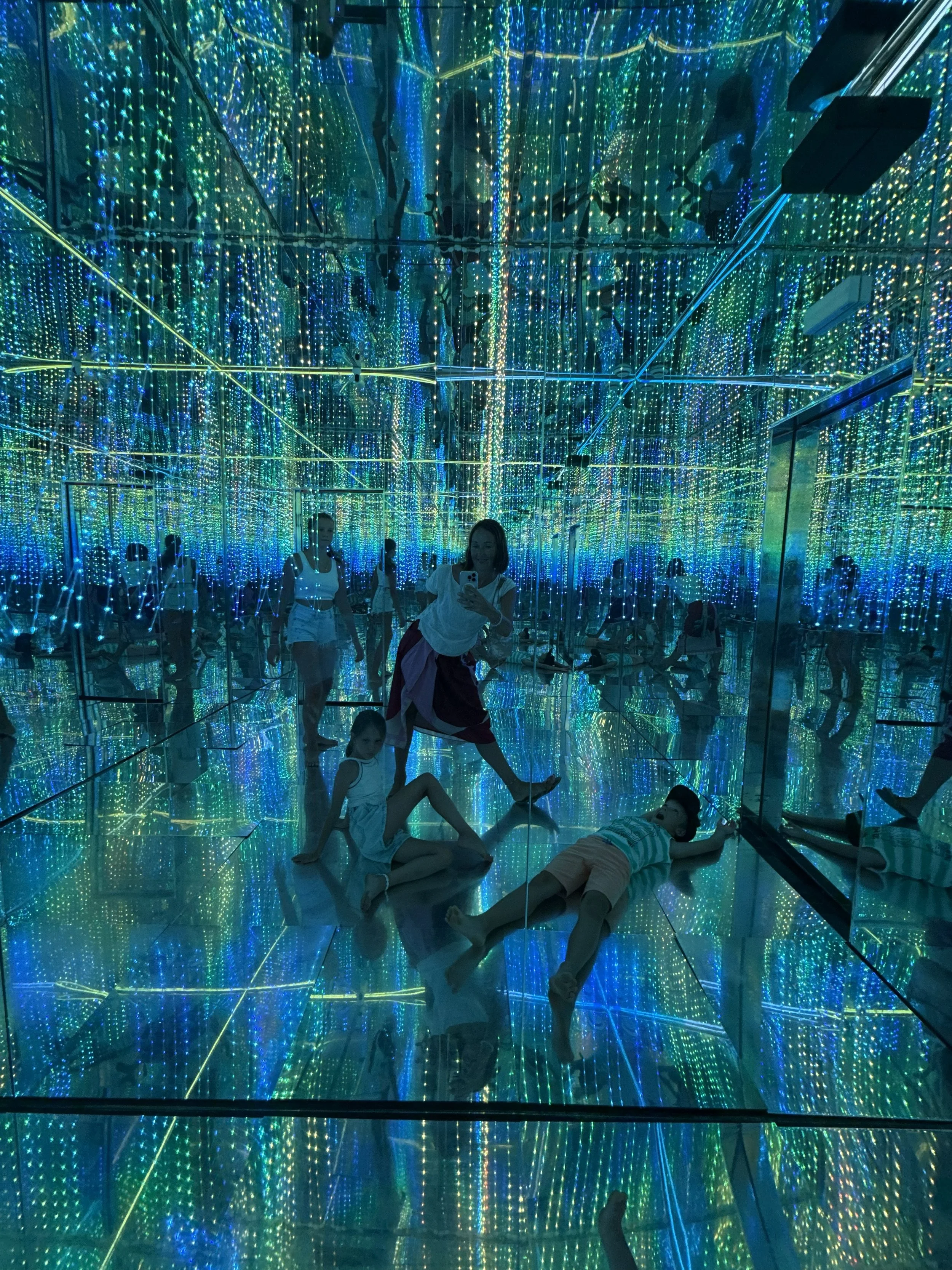 People inside an interactive mirror maze with colorful LED lights reflecting on mirrored walls and floor.