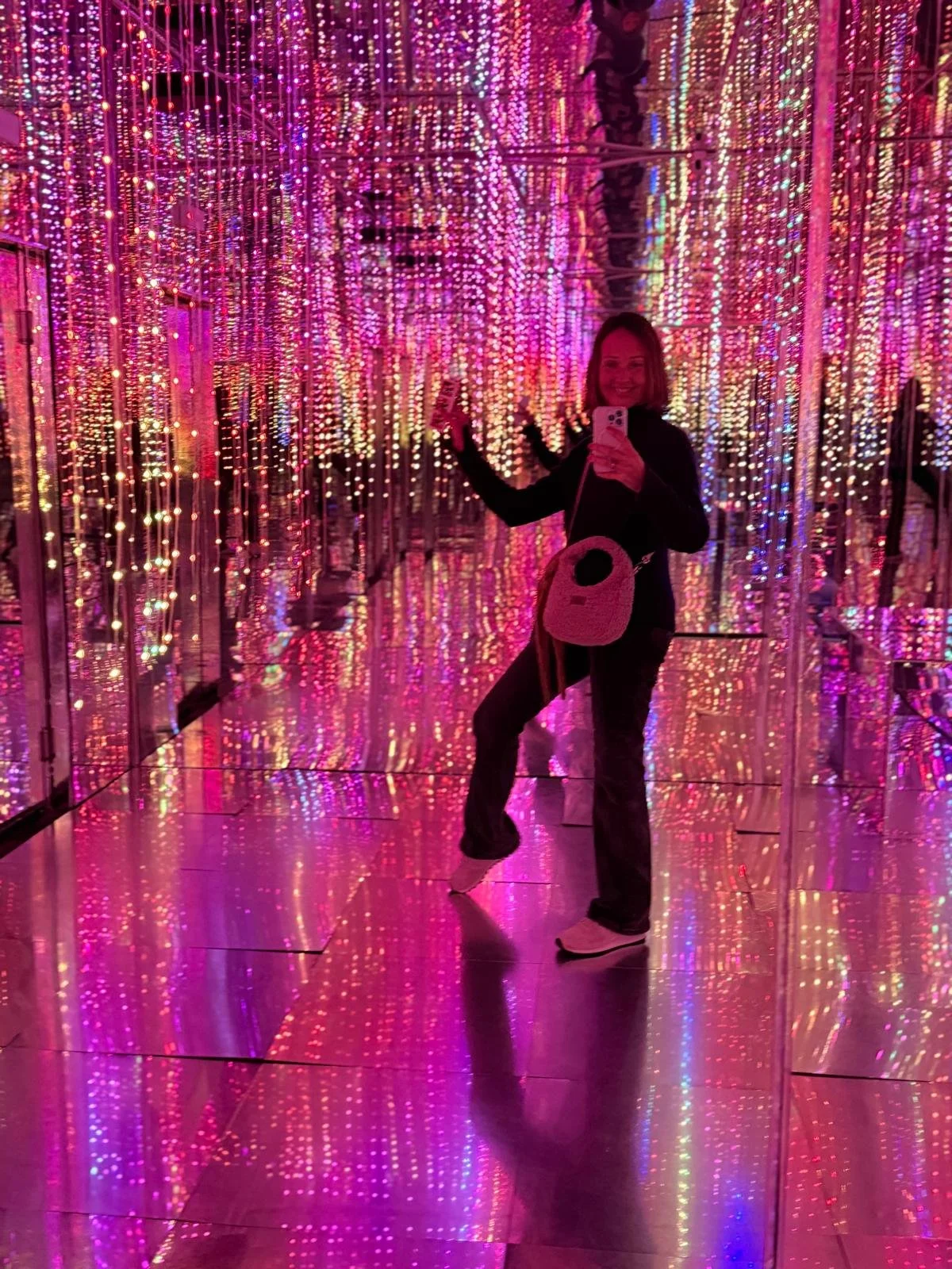 A woman taking a selfie in a room with walls and ceiling covered in pink, purple, and blue LED light strips, creating a colorful, luminous environment.