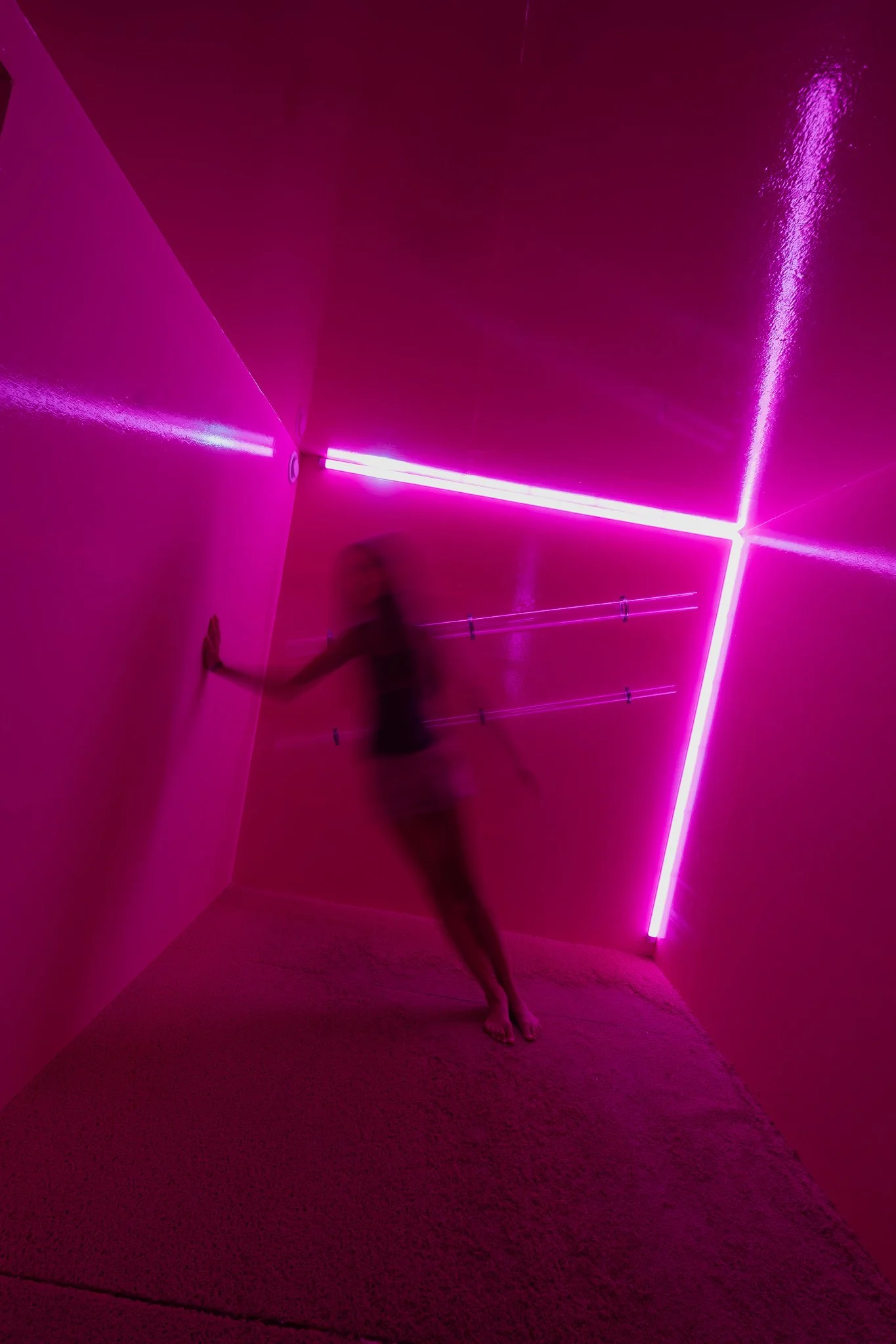 Person standing in a pink-lit room with neon light art on the wall, blurry due to motion.