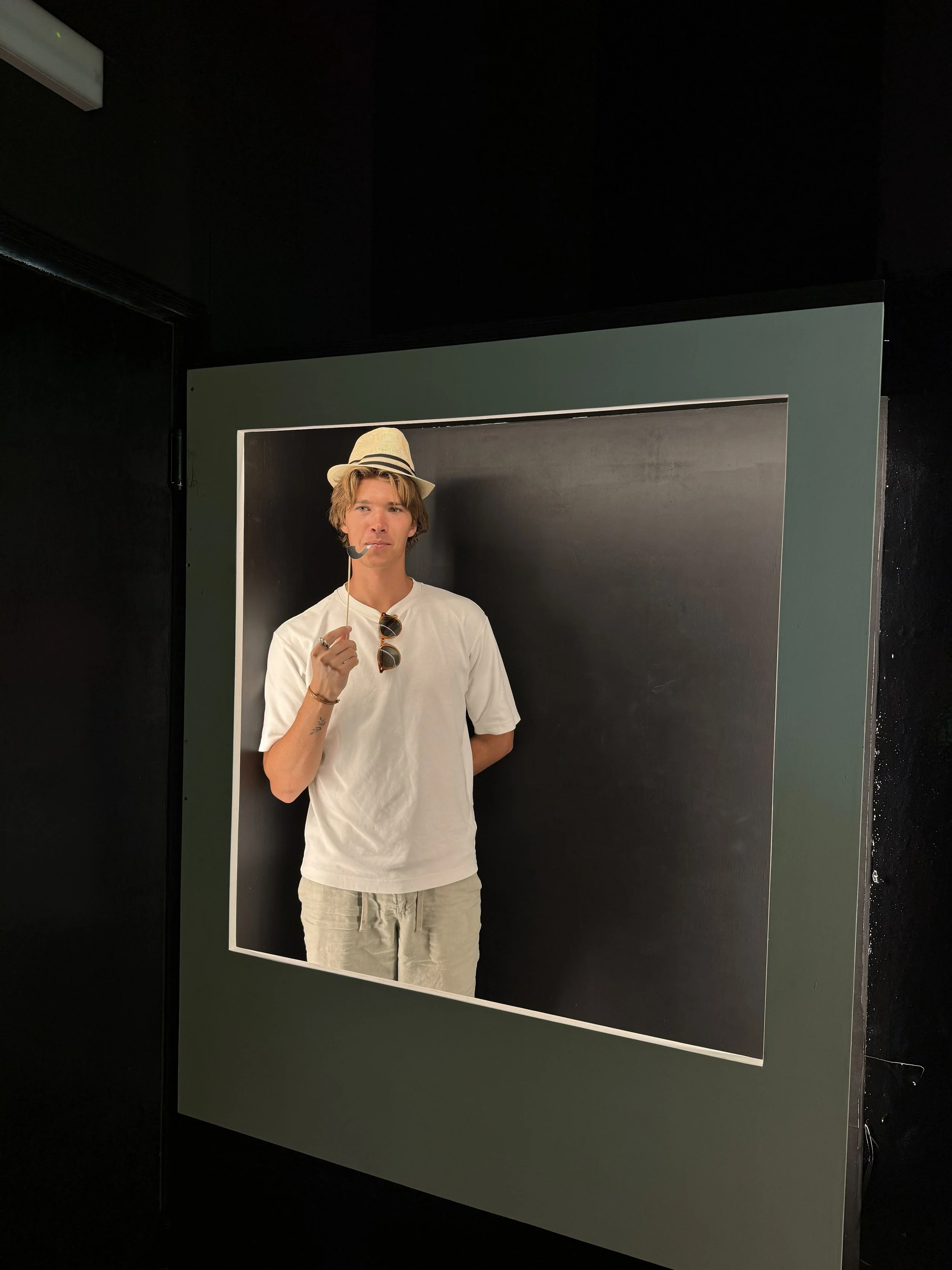 Young man wearing a straw hat, sunglasses hanging from shirt, holding a pipe, standing in a black photo booth with a dark background.