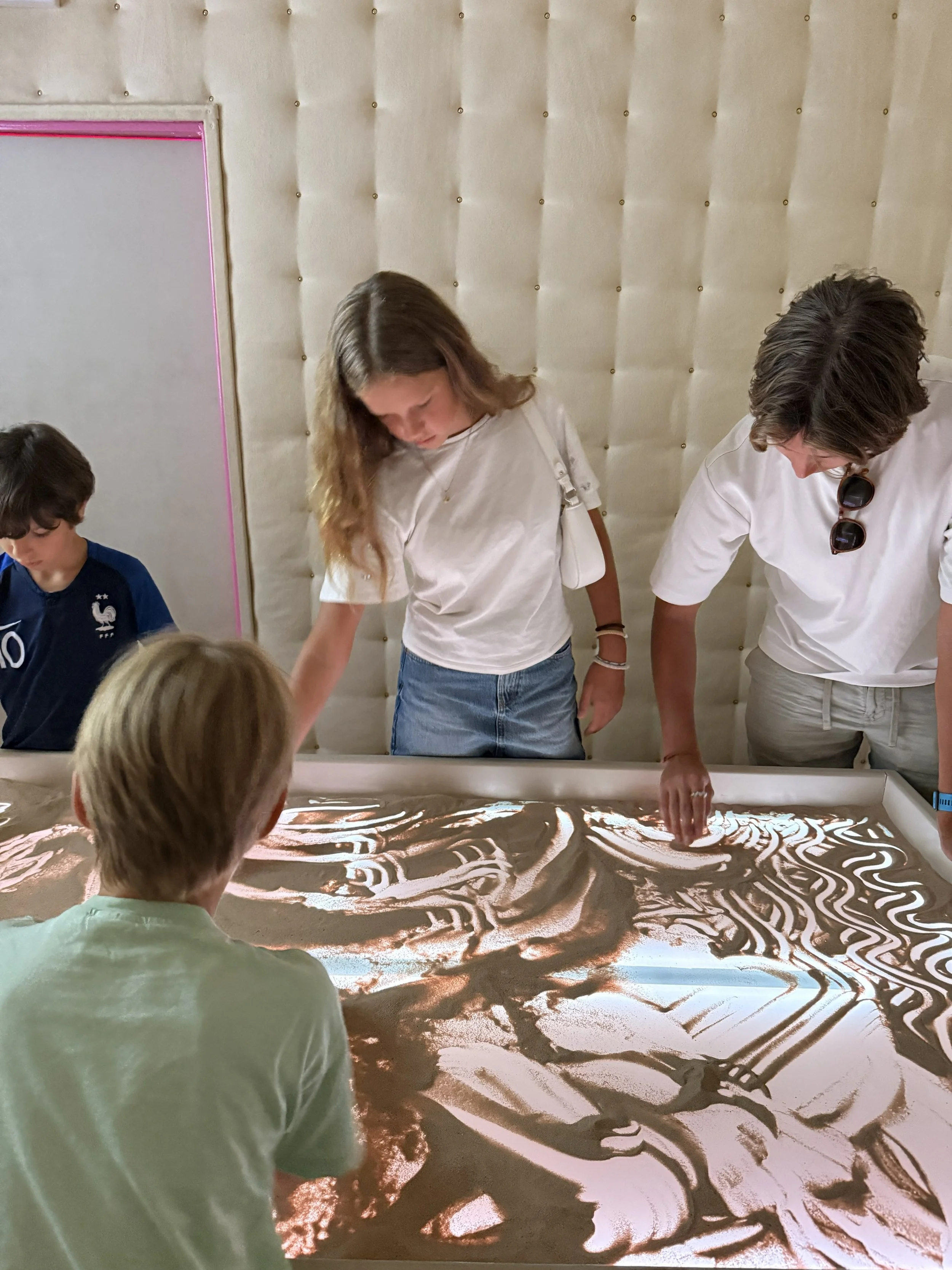 Children and a woman are gathered around an illuminated sand art panel creating artistic designs with sand.