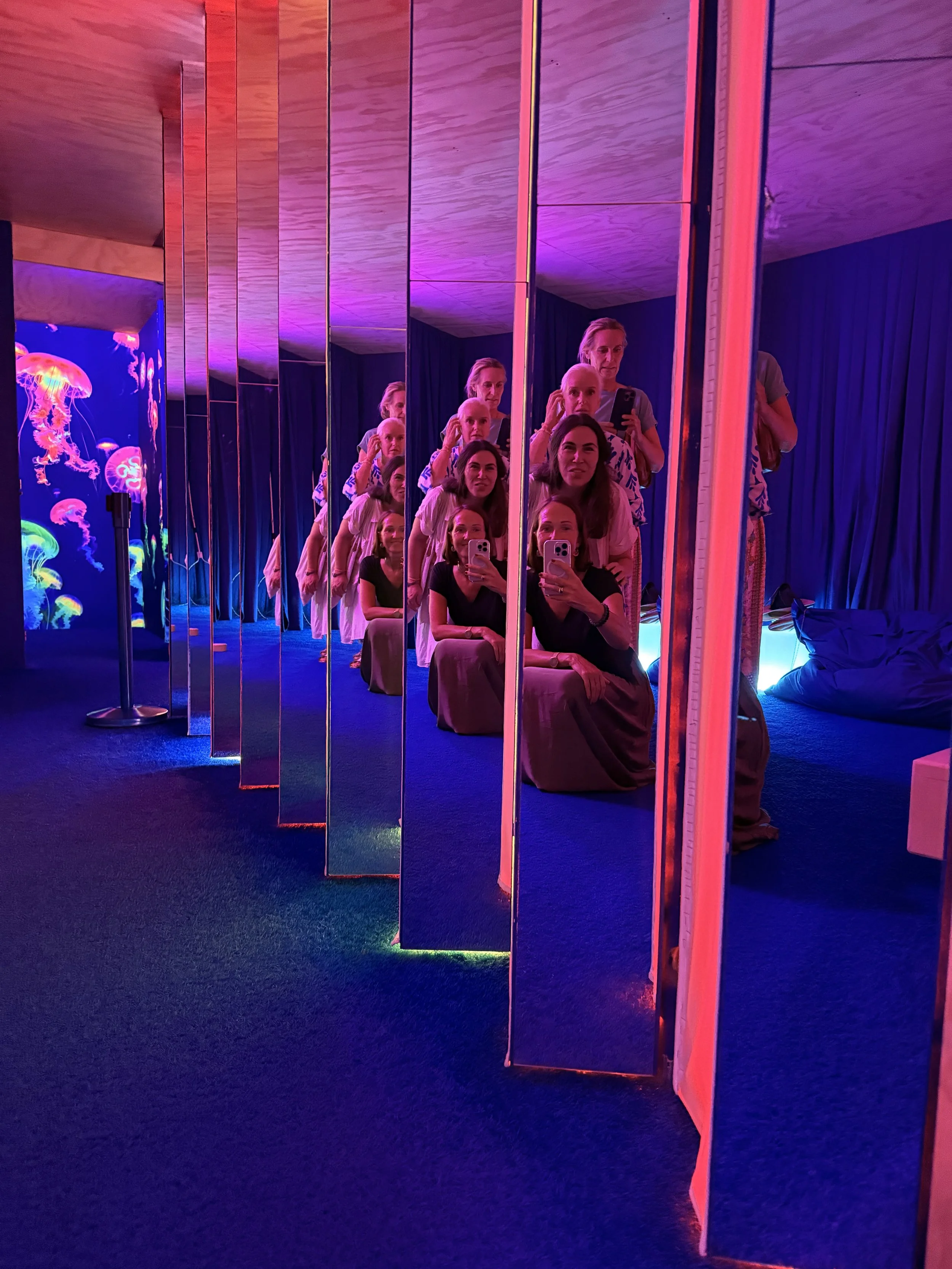 Multiple women and a girl sitting and kneeling in front of mirrored panels, taking selfies in a colorful, neon-lit room with jellyfish images on a digital screen in the background.
