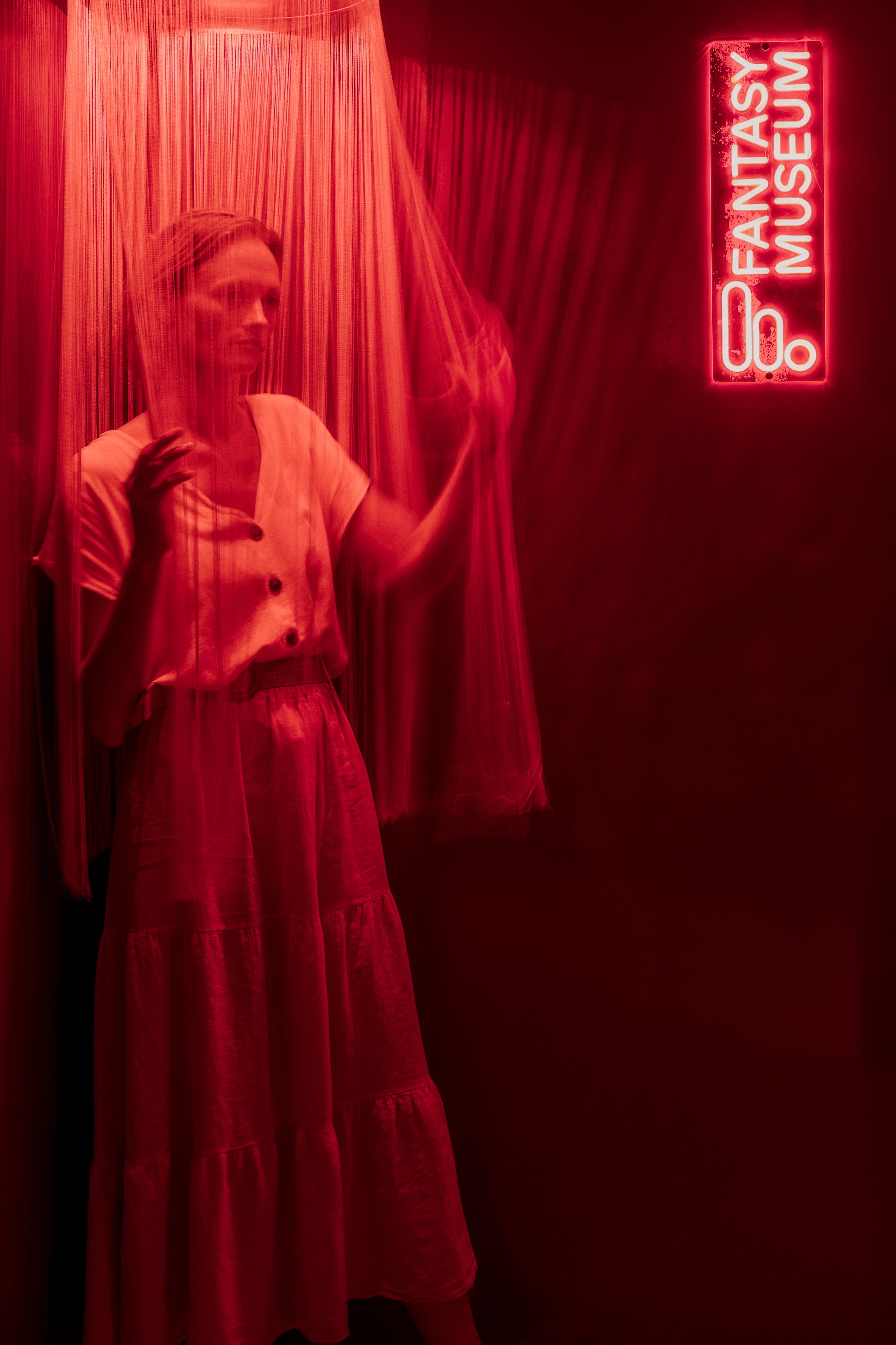 A woman standing behind a red curtain, touching it with one hand, inside the Fantasy Museum which is marked by a neon sign on the wall.