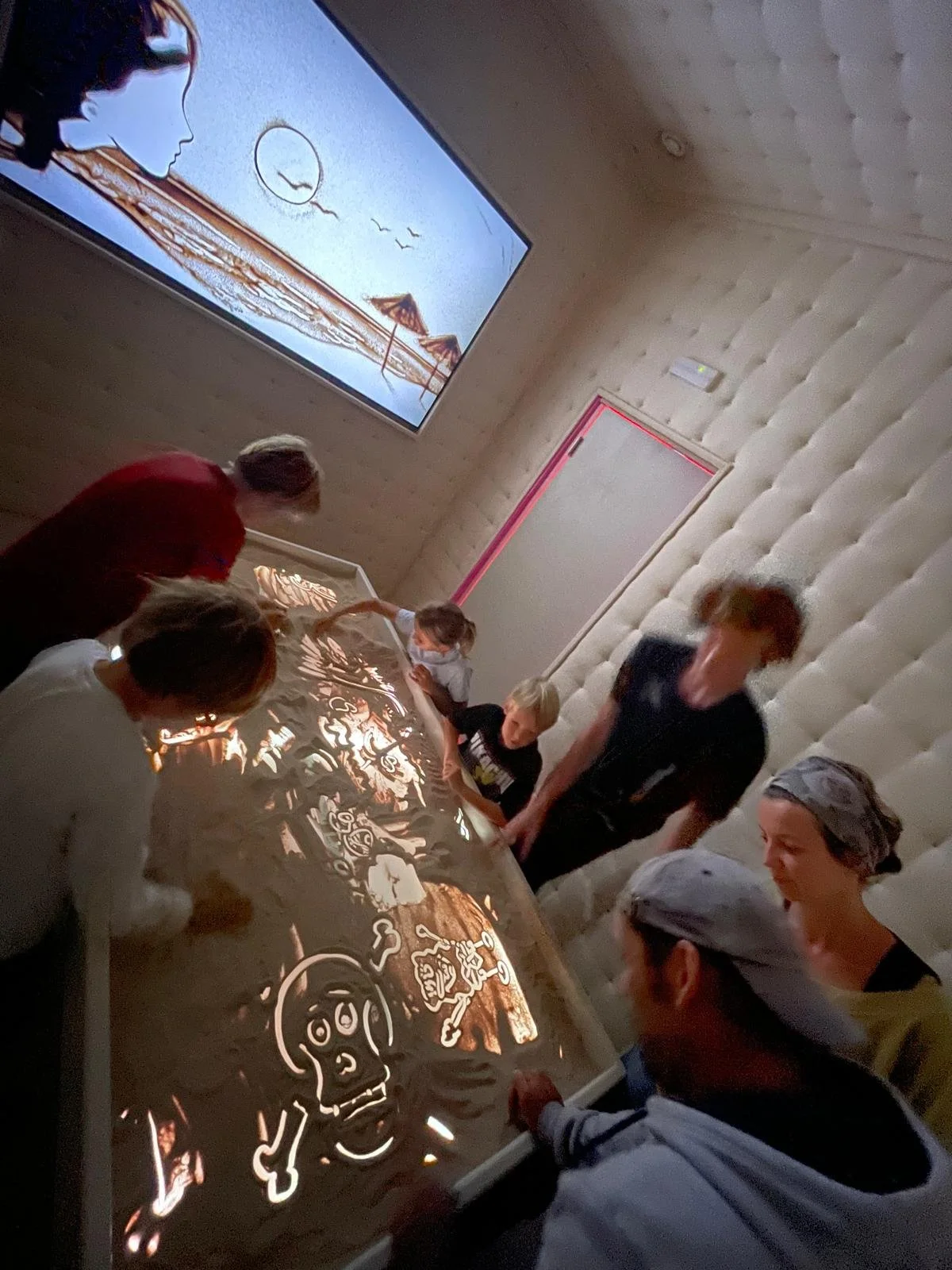 People gathered around a light-up table drawing with illuminated sand or light-sensitive material while a large screen displays beach-themed artwork in the background.