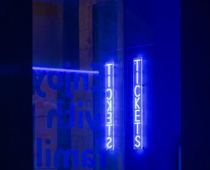 Neon blue sign reading 'TICKETS' and another one reading 'TICKETS' in reflections on a glass wall.