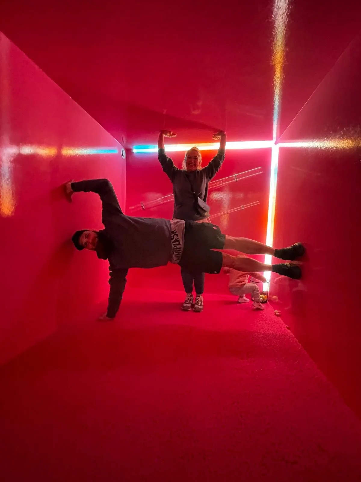Two people in a narrow, red-lit tunnel, one bending sideways with their hand on the floor, the other standing and raising their arms above their head.