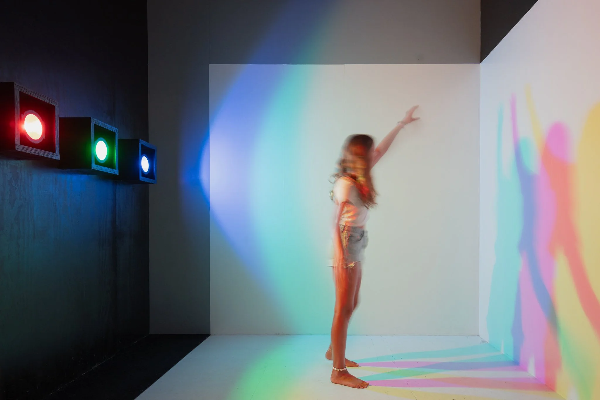 A woman standing with her arm raised in a light-colored room with colorful lighting effects projected onto the wall and floor, and three colored lights (red, green, blue) on a black wall to her left.