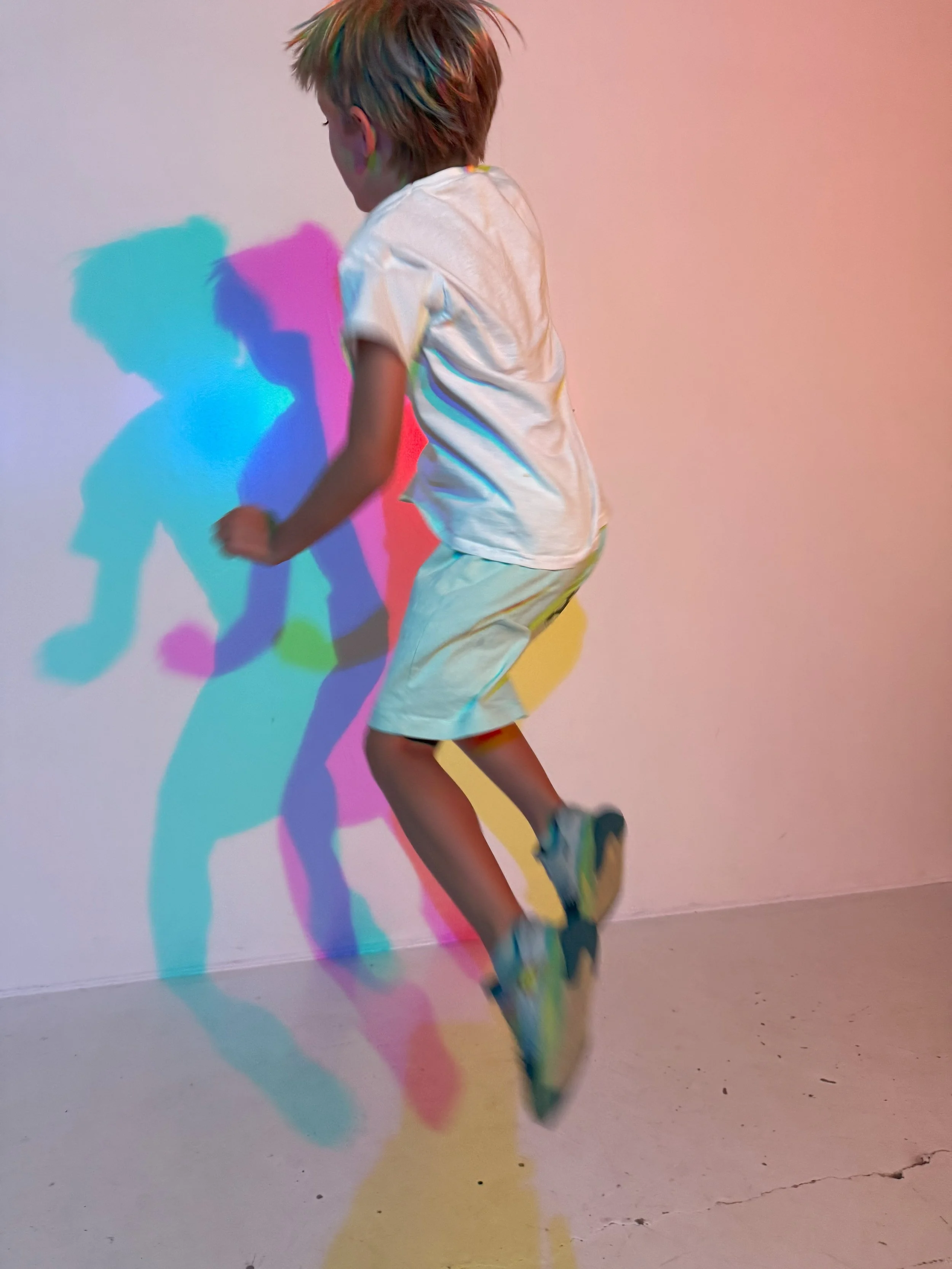 A young boy jumping with colorful shadow projections on the wall behind him, wearing a white t-shirt, light-colored shorts, and sneakers.