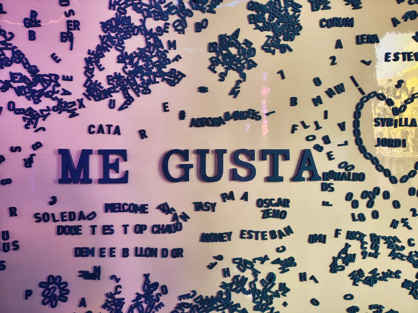 Decorative wall with scattered blue letter and number magnets, with large central text saying "ME GUSTA" and hashtags such as "#AURORA#ANGELS" among other words.