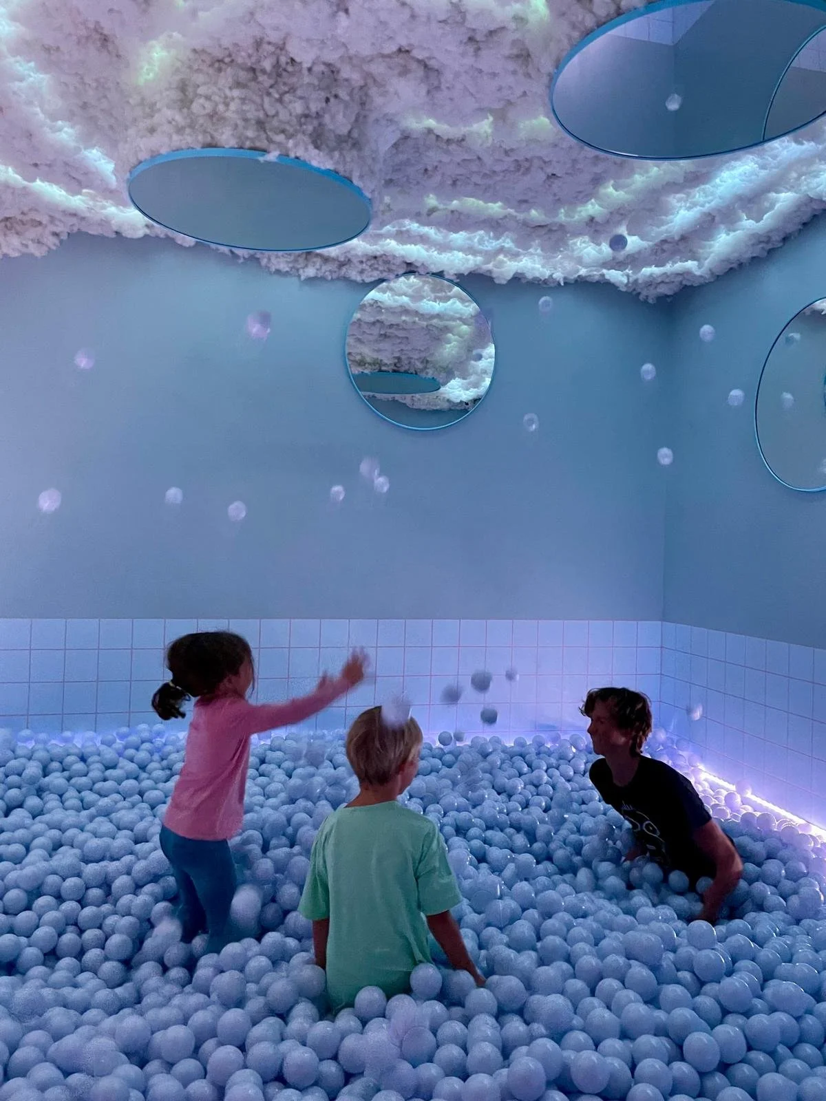 Three children playing in a ball pit with a cloud-like ceiling and round mirrors on the walls, illuminated with blue lighting.