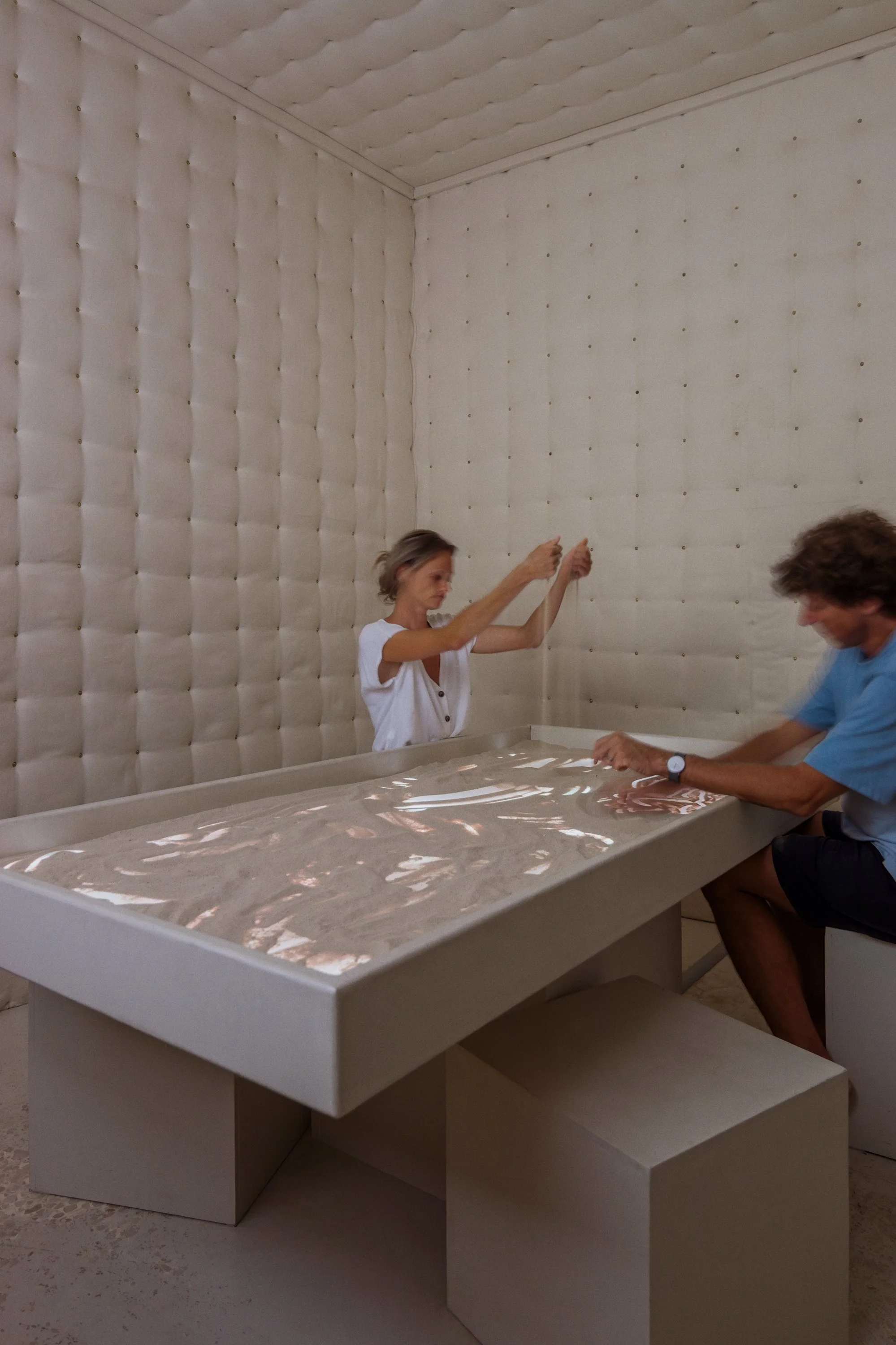 Two people sitting at a rectangular table with a reflective surface, inside a padded room with quilted, beige walls.