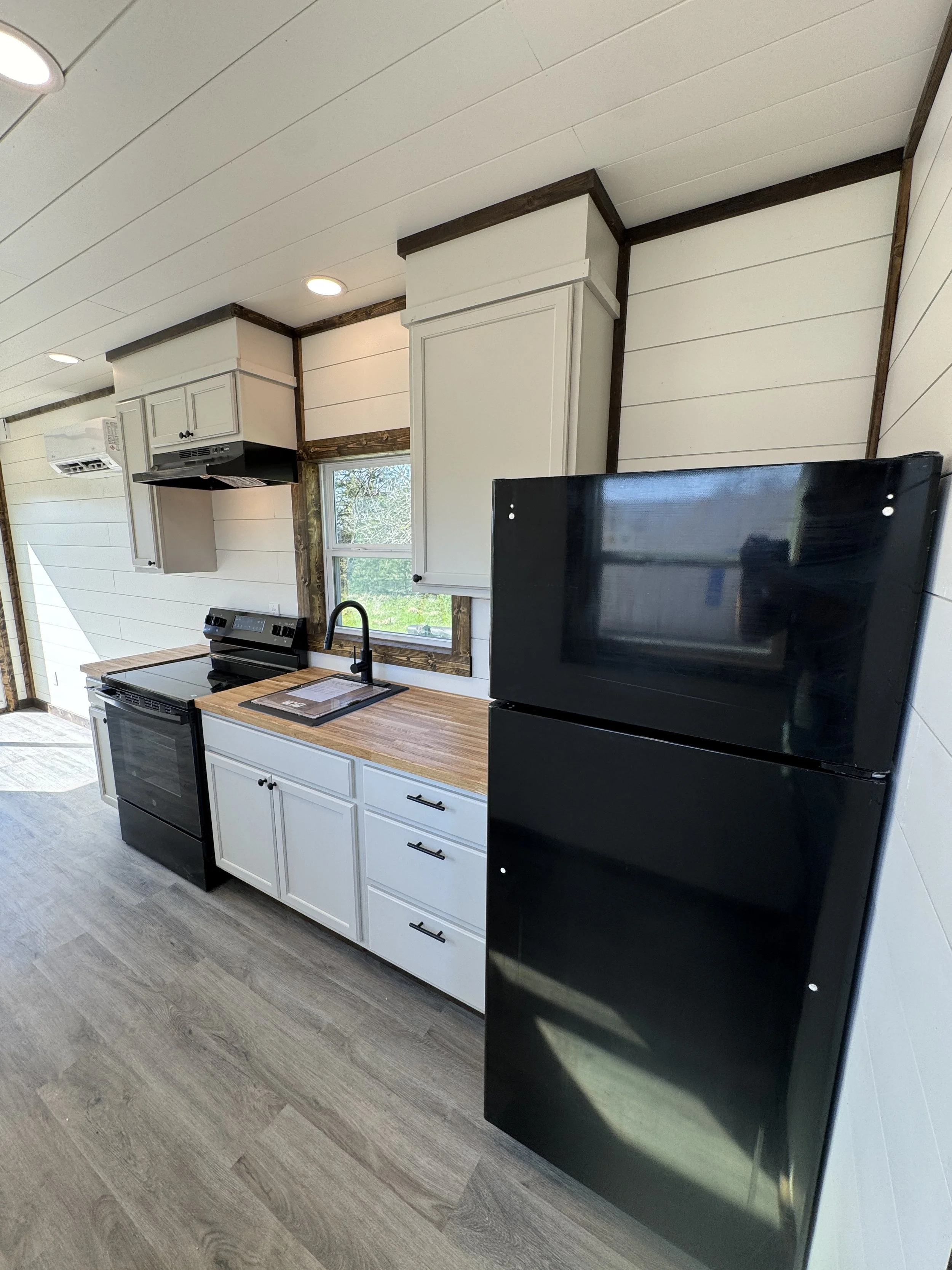 Modern kitchen interior in turnkey shipping container home in Paris Texas with white shaker cabinets, black appliances, wood butcher block countertops, black gooseneck faucet, and large window