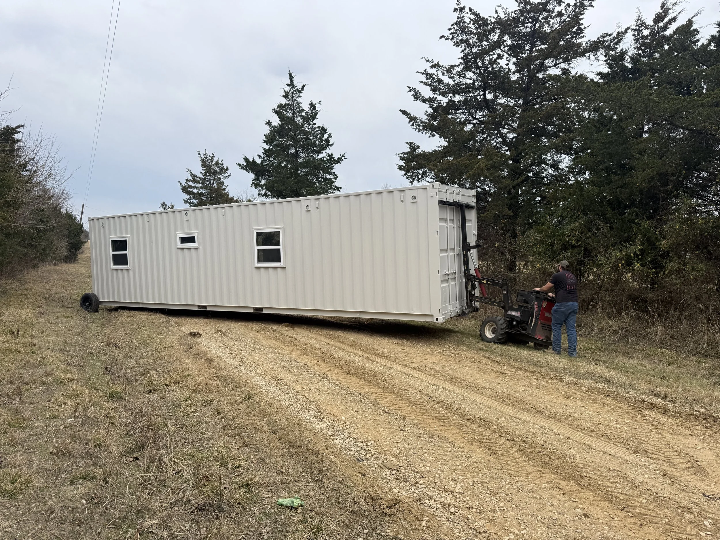 Shipping container home being professionally delivered and positioned on site in Paris Texas using telehandler forklift with worker operating equipment on grassy lot with trees in background