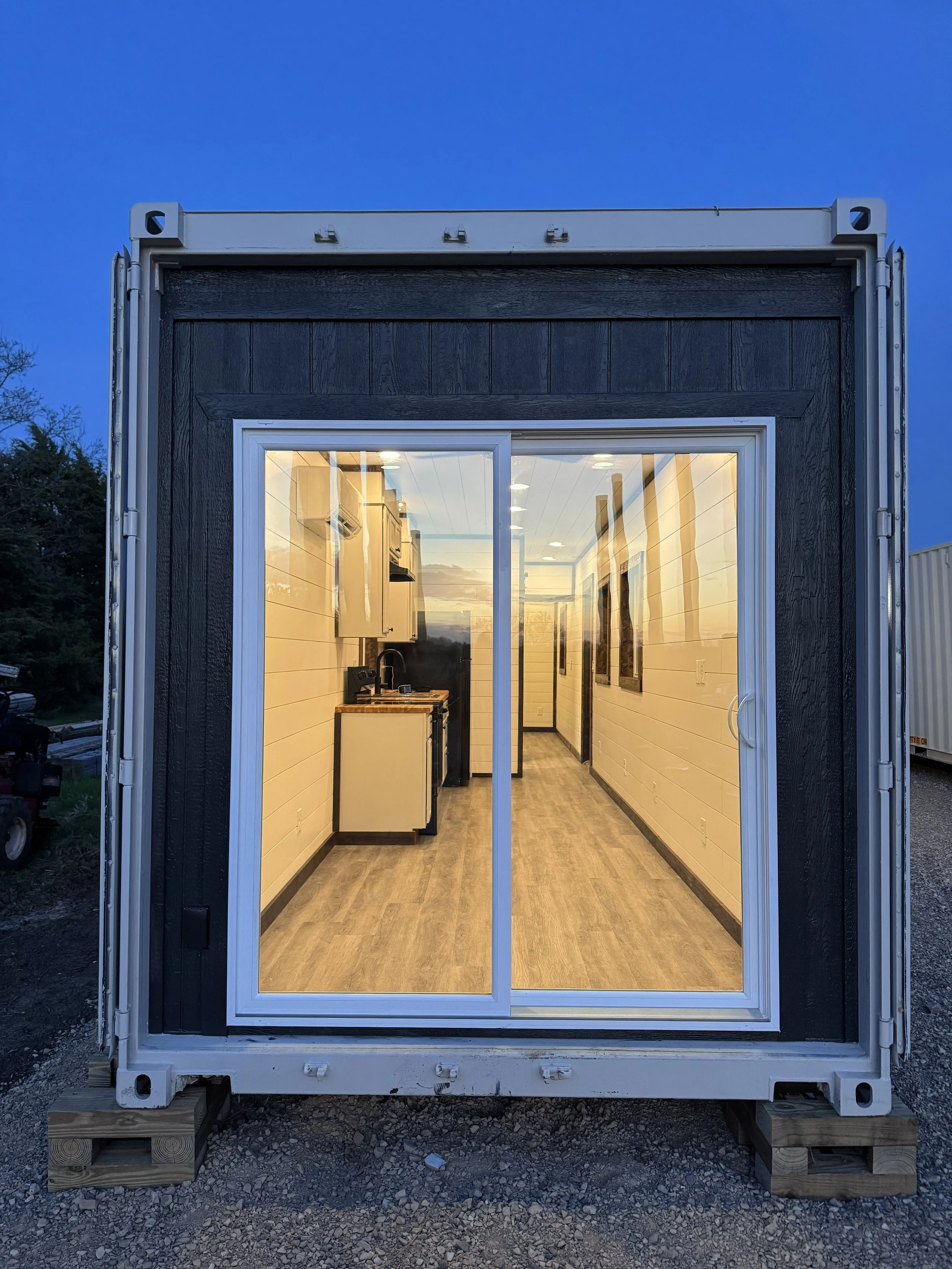 View of a tiny house with large sliding glass doors revealing a brightly lit interior with a small kitchenette and artwork on the walls, set on a gravel lot during dusk.
