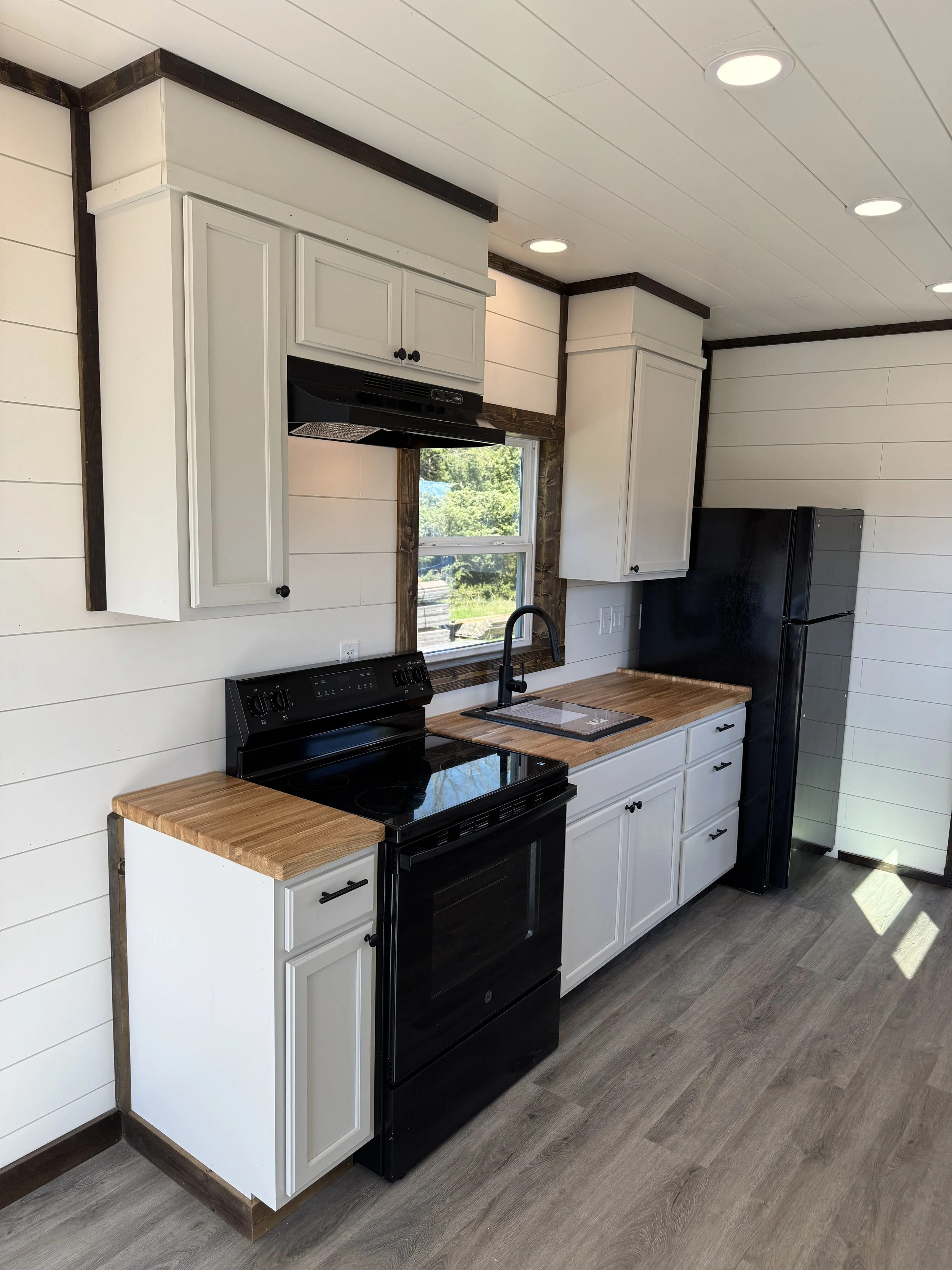 Modern kitchen interior of turnkey shipping container home in Paris Texas with white shaker cabinets, black appliances, wood butcher block countertops, large window, and shiplap walls