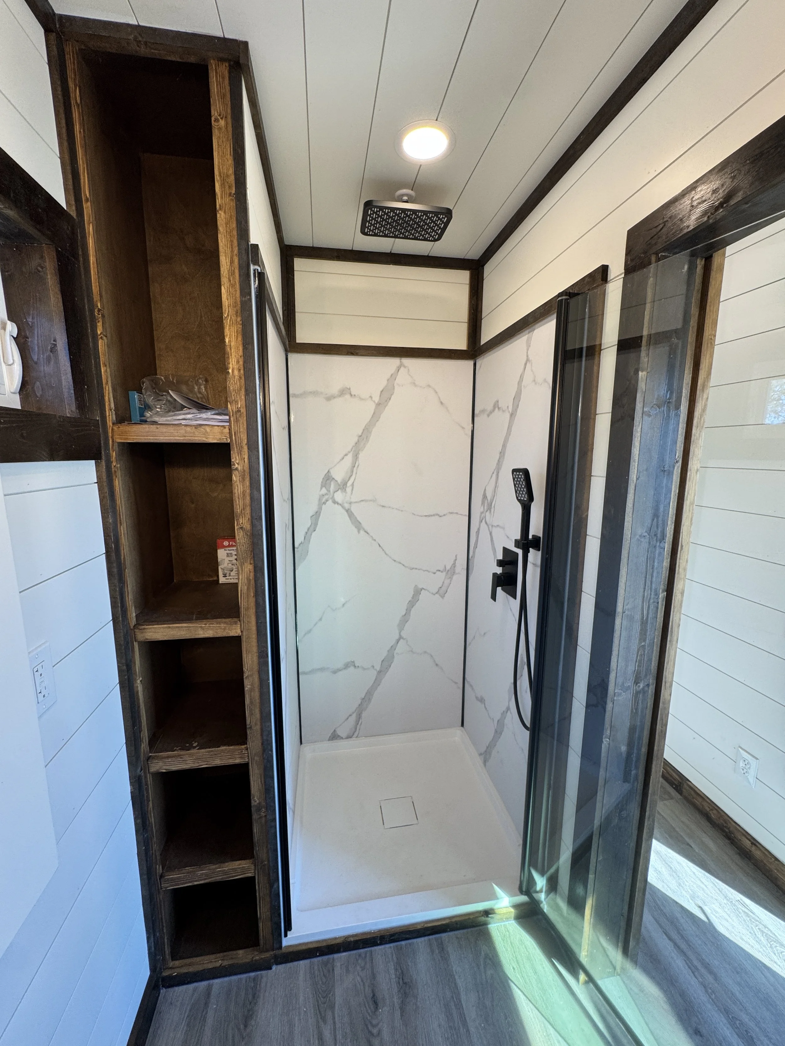 Modern walk-in shower in turnkey shipping container home in Paris Texas with marble-patterned walls, black matte fixtures, glass enclosure, rain shower head, and built-in wooden shelving