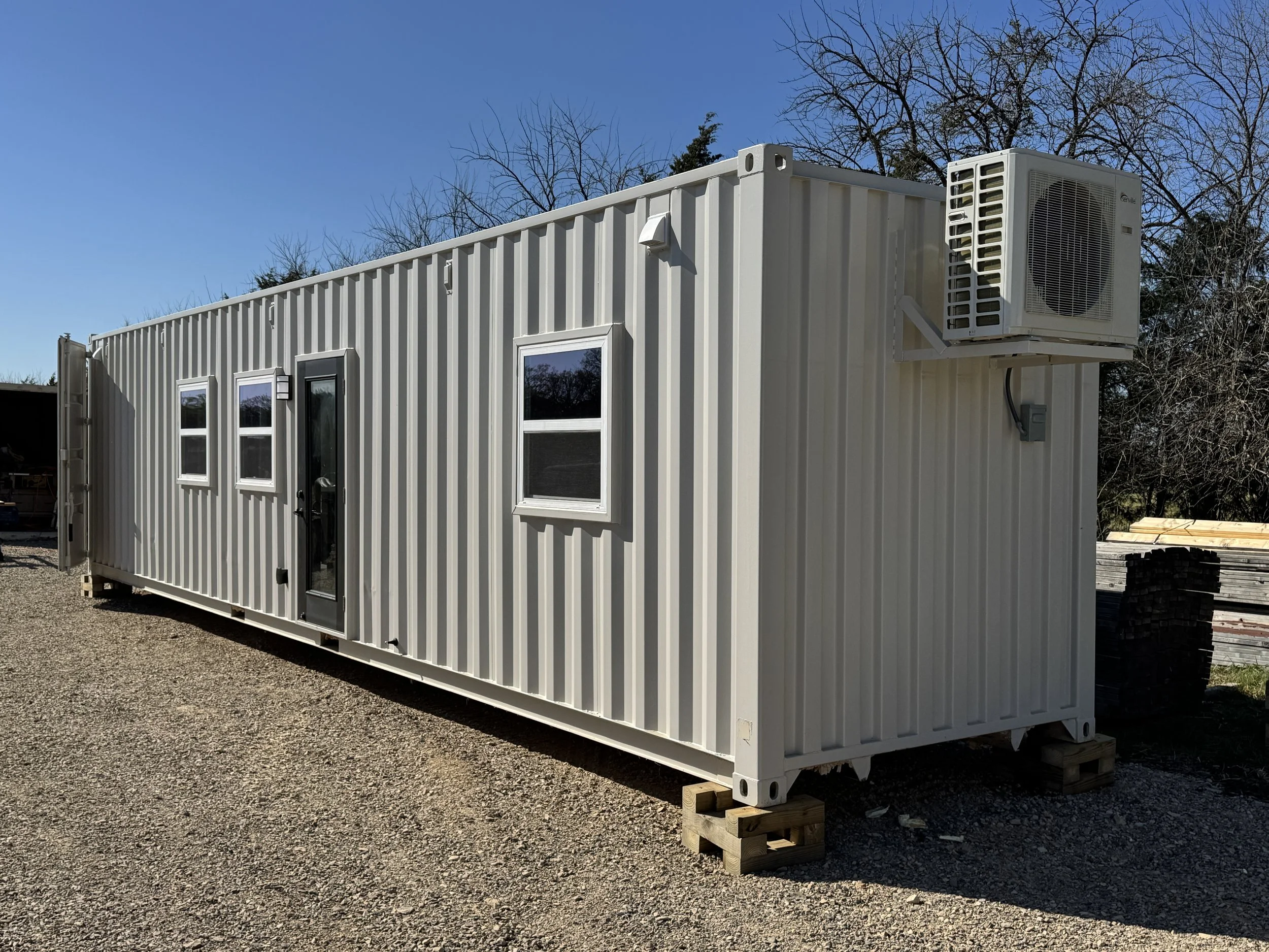 Turnkey shipping container home exterior in Paris Texas with large sliding glass patio doors, white steel siding, multiple windows, and wall-mounted mini-split air conditioner on a sunny day with gravel lot