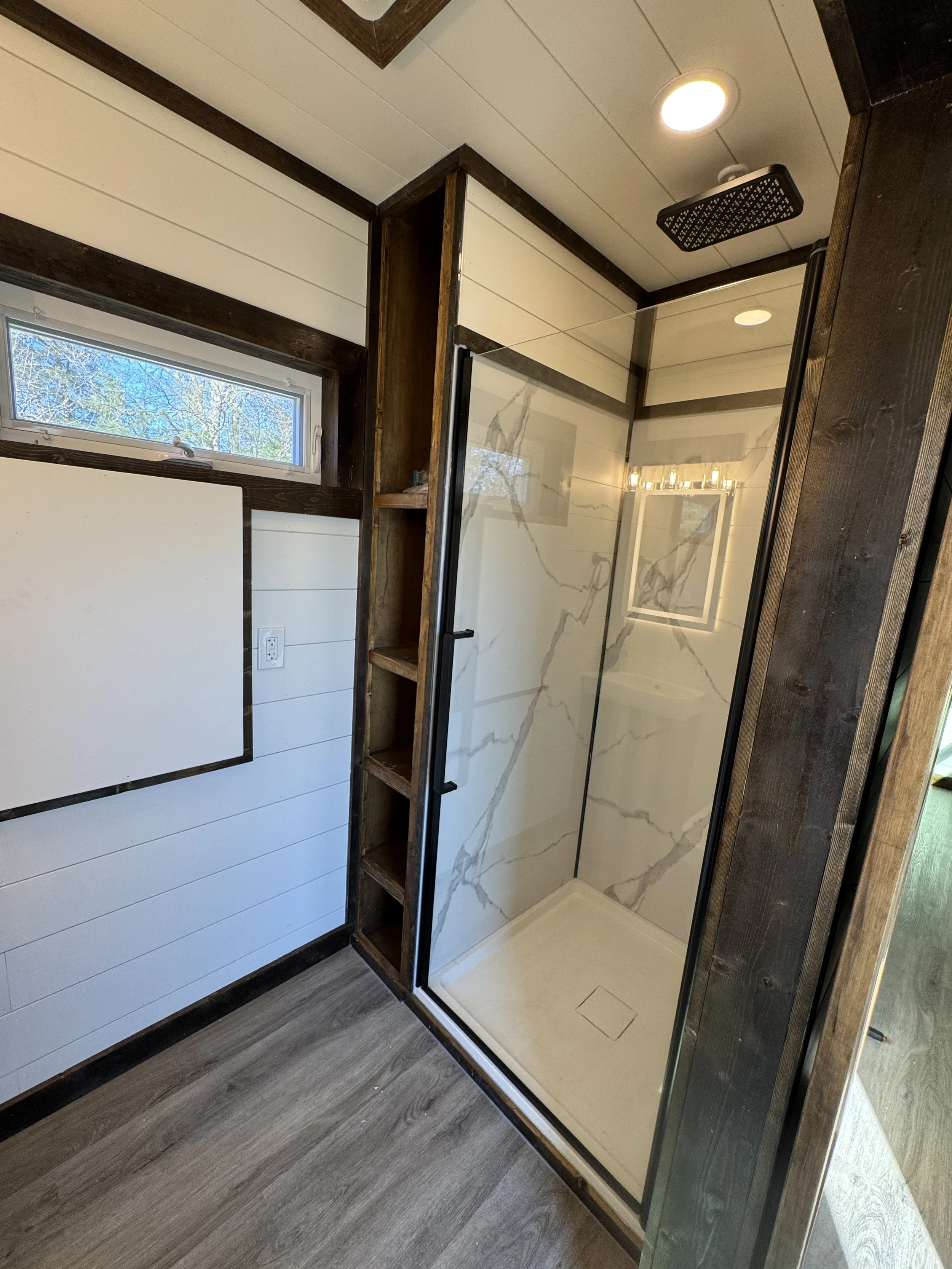 Modern bathroom interior in turnkey shipping container home in Paris Texas with walk-in shower featuring glass door and marble-patterned walls, white shiplap, dark wood accents, large window, and built-in shelving