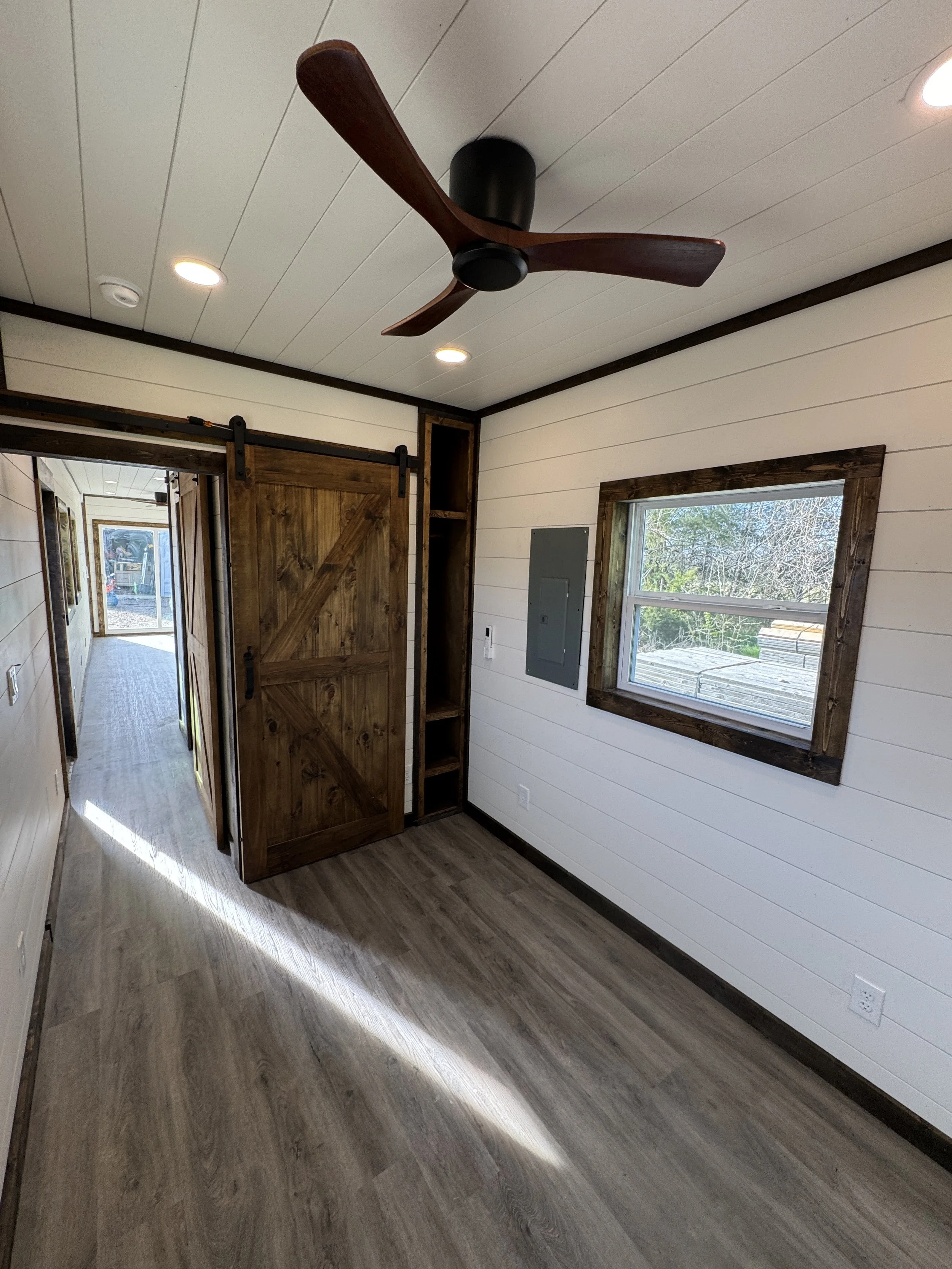 Modern shipping container home interior in Paris Texas with white shiplap walls, dark wood sliding barn door, ceiling fan, recessed lighting, and warm natural light from the large window