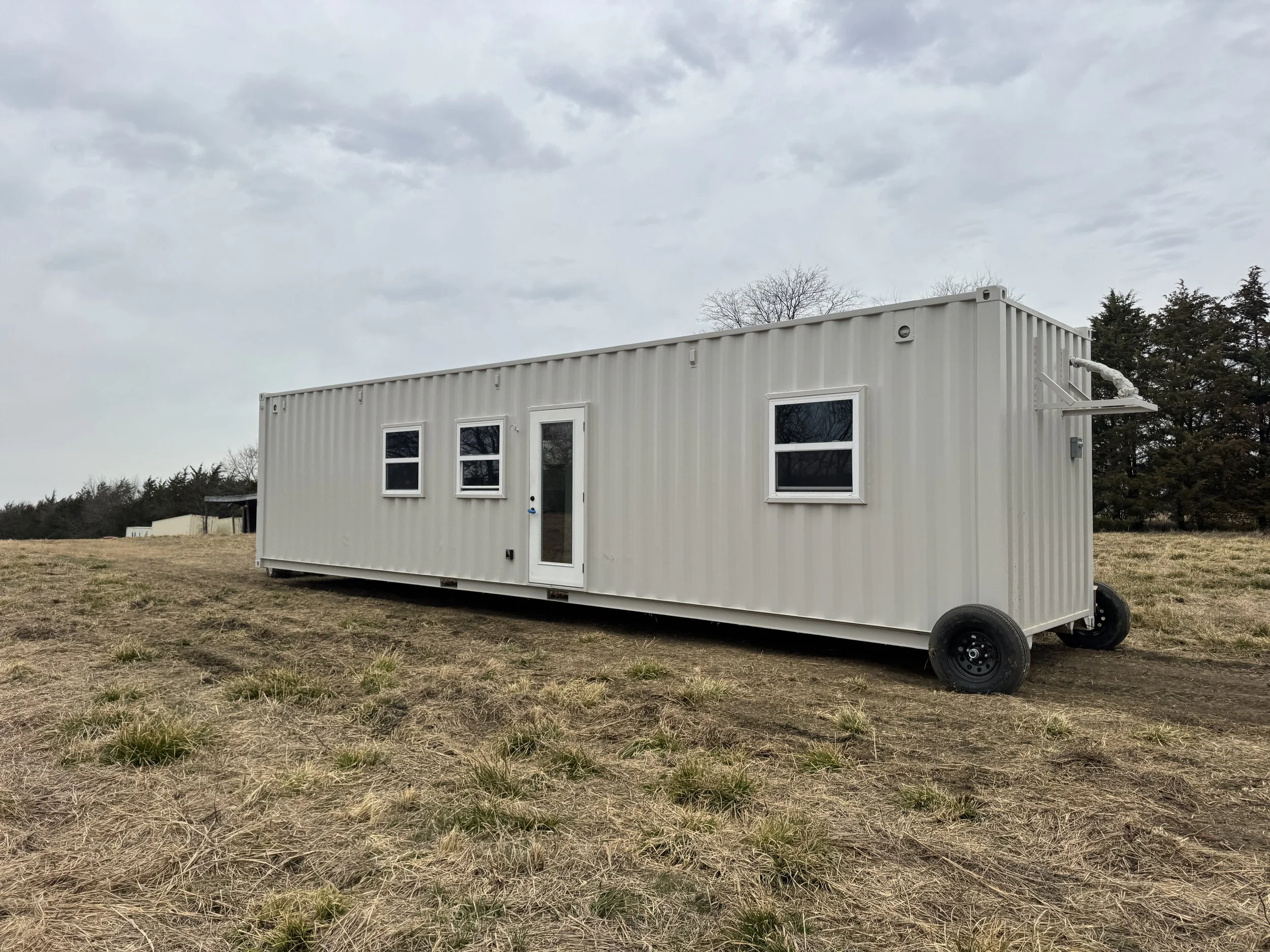 Turnkey shipping container home exterior in grassy field in Paris Texas with white steel siding, windows, entry door, and wheels visible under cloudy sky